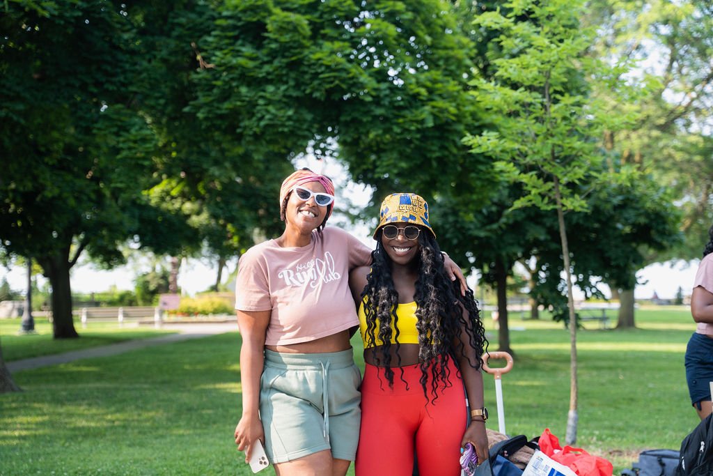 Two women smiling and posing outdoors in a park, standing close together with one woman's arm around the other. They are dressed in colorful casual summer outfits, with sunglasses and hats. Trees and greenery are visible in the background.