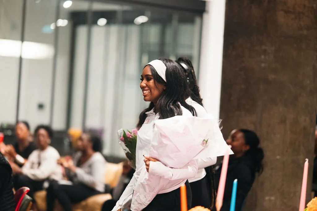 A woman with a white headband and long wavy black hair holding a bouquet of flowers, smiling at an indoor event with seated people in the background.