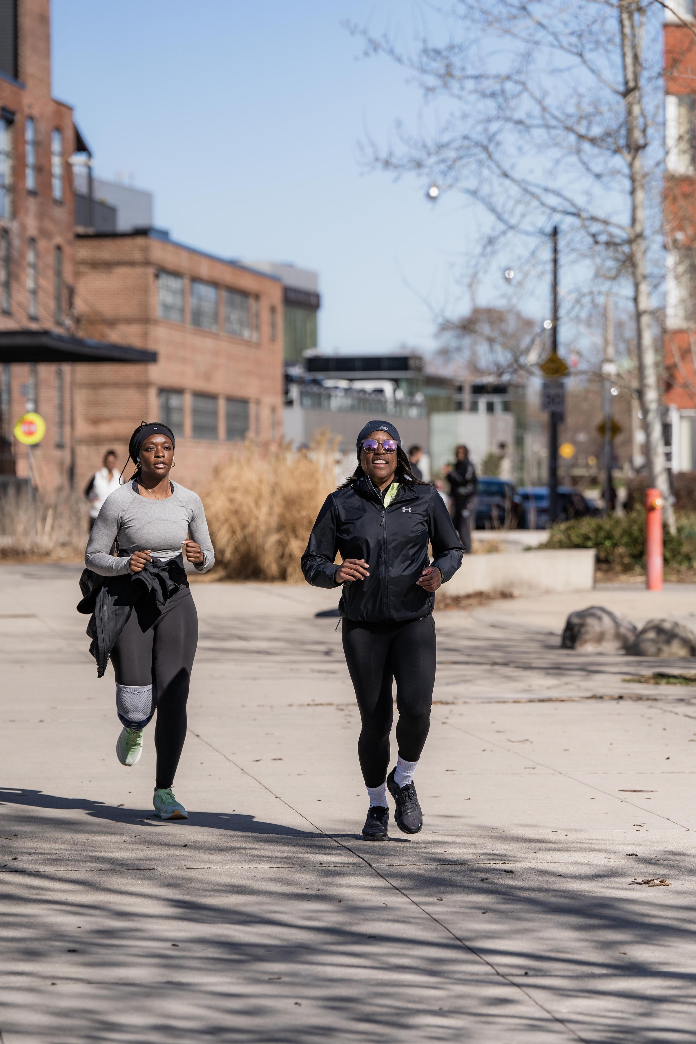 Two women jogging together outdoors on a sunny day with buildings and trees in the background.