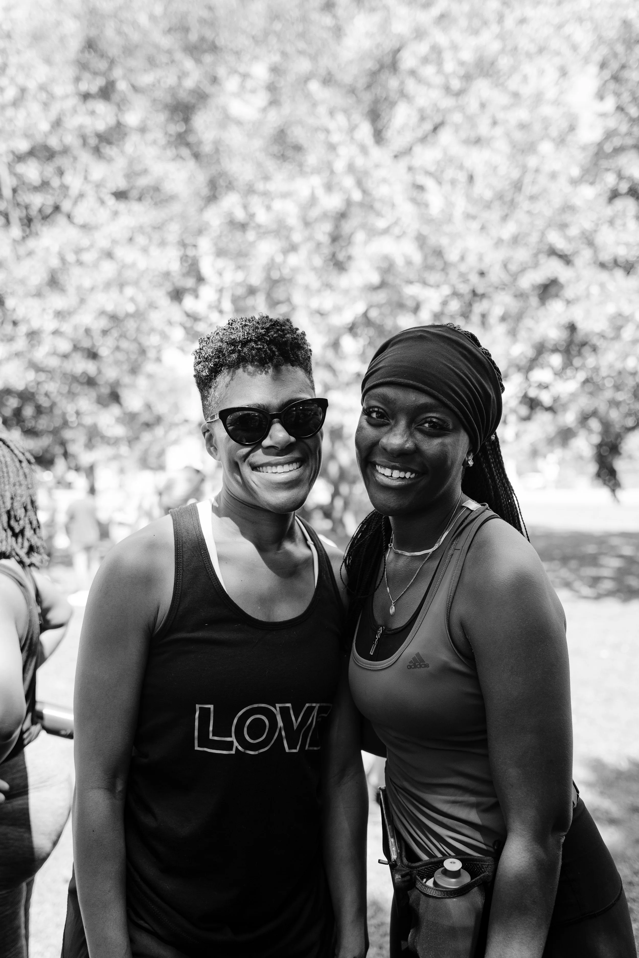 Two women smiling outdoors, one wearing sunglasses and a sleeveless top with the word "LOVE," the other wearing a headscarf and athletic clothing, with trees in the background.