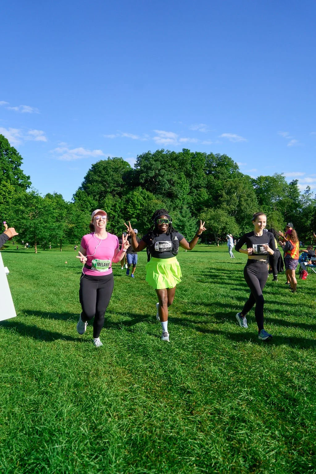 Three women running in a park during a race or marathon, smiling and making peace signs, with other participants and green trees in the background on a sunny day.