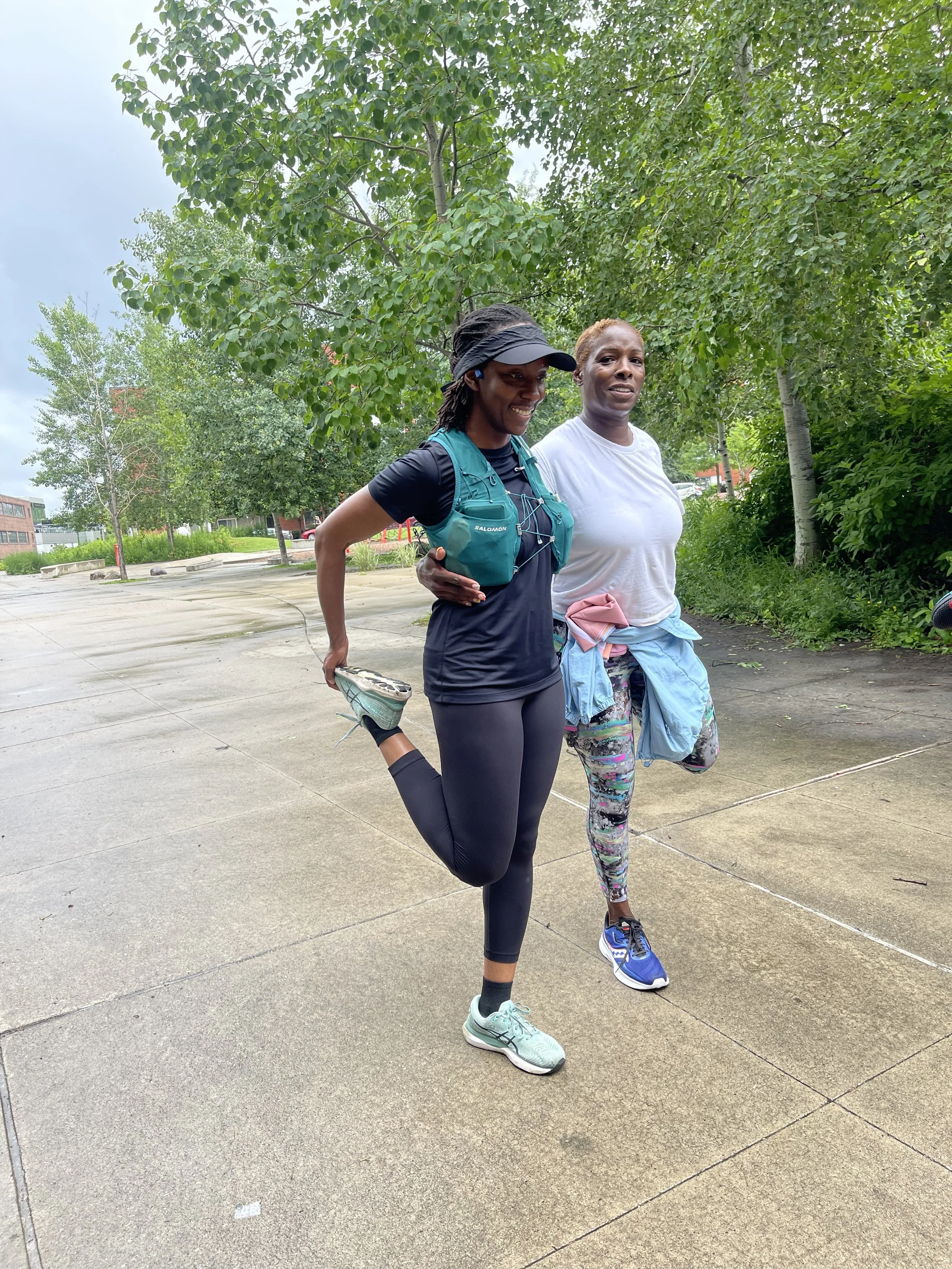 Two women are outdoors on a sidewalk, one is stretching her leg behind her while holding her ankle, and both are smiling. The woman on the left is wearing a black cap, black athletic outfit, and teal running shoes. The woman on the right is wearing a