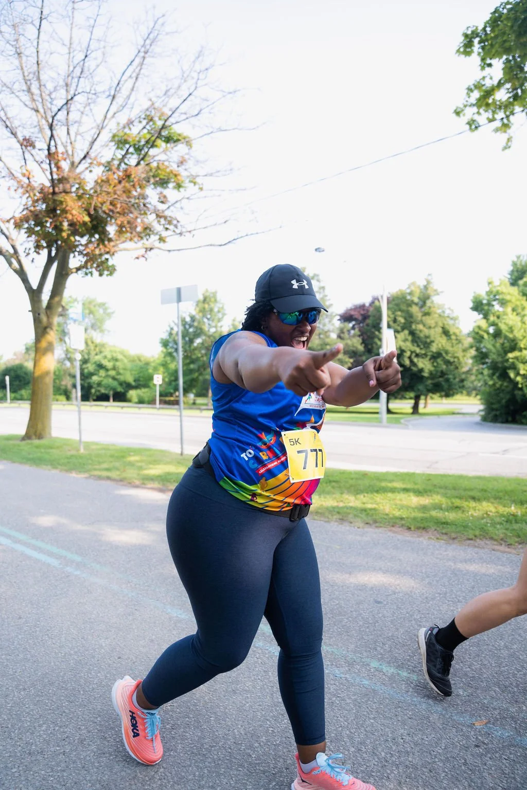 A woman running in a race, wearing sunglasses, a black cap, a blue and rainbow-colored athletic shirt, black leggings, and pink running shoes, smiling and pointing as she crosses the finish line.