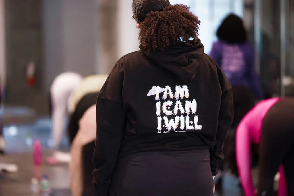 Back view of a woman in a black hoodie with a motivational message, standing among people in a gym or fitness class setting.