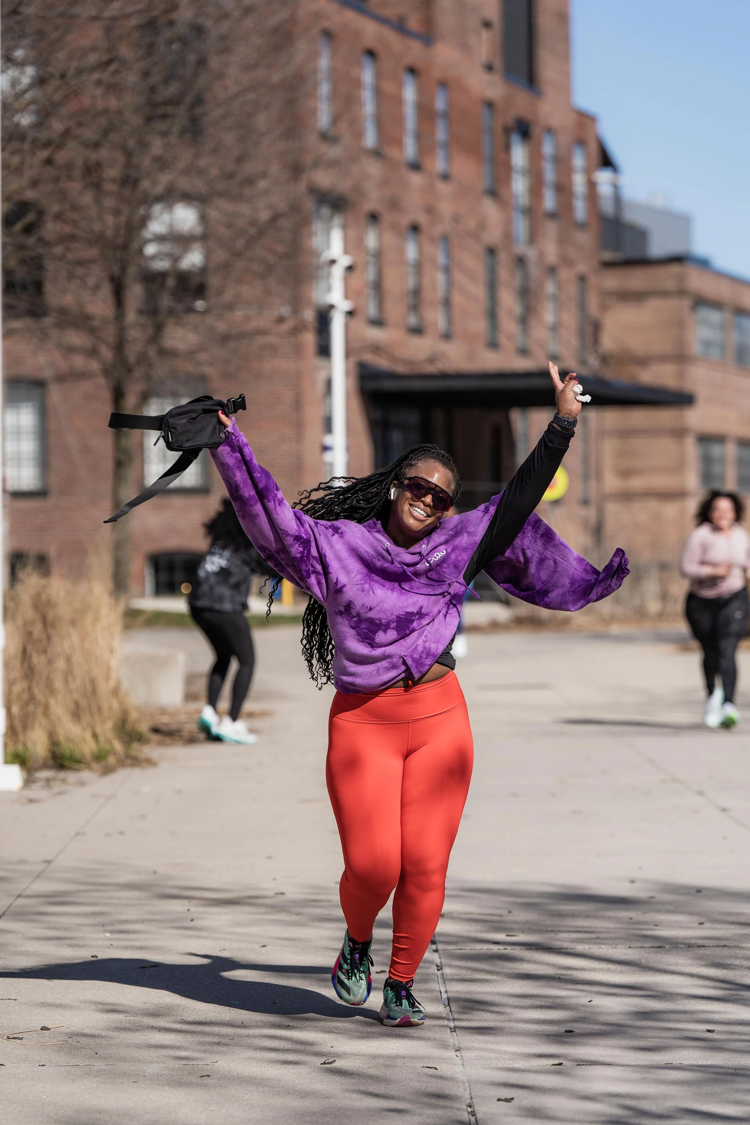 A woman smiling and running outdoors during daytime, wearing a purple tie-dye hoodie, red leggings, and sunglasses, with her arms raised in celebration.