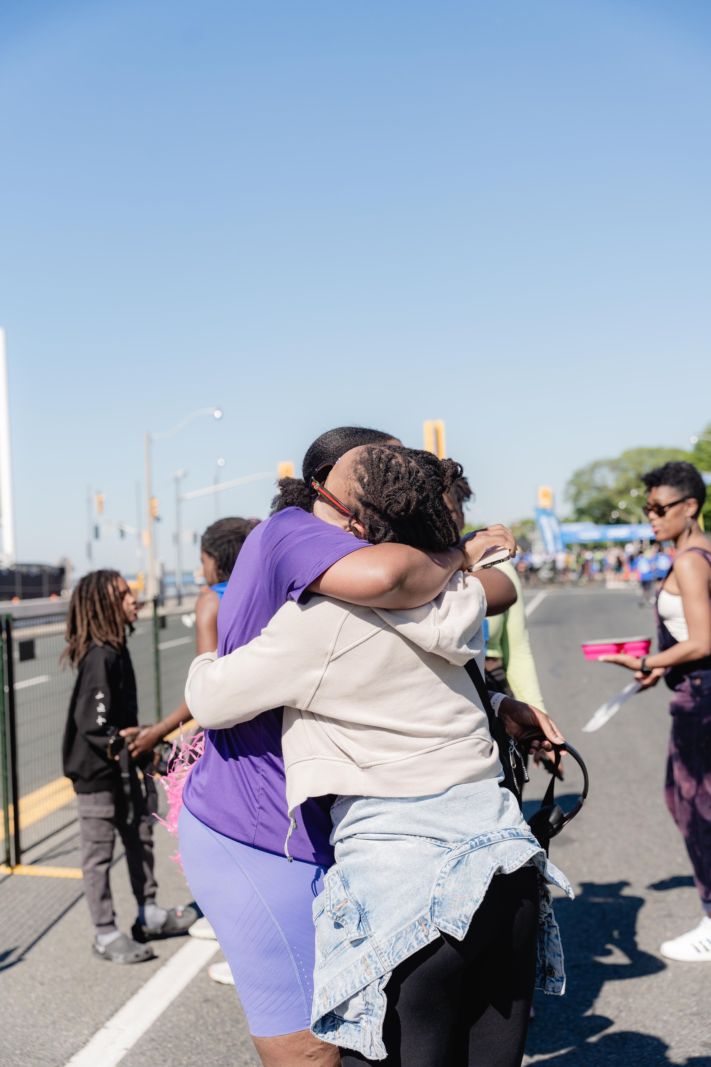 Two people hugging each other at an outdoor event on a sunny day, with other people and a street in the background.