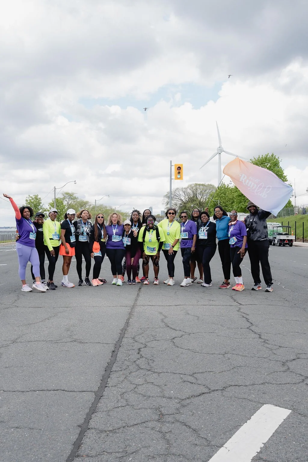Group of women and one man participating in a marathon or run, standing on a street with cloudy sky, wind turbine, and trees in the background, some holding a flag that says 'RUN Club'.