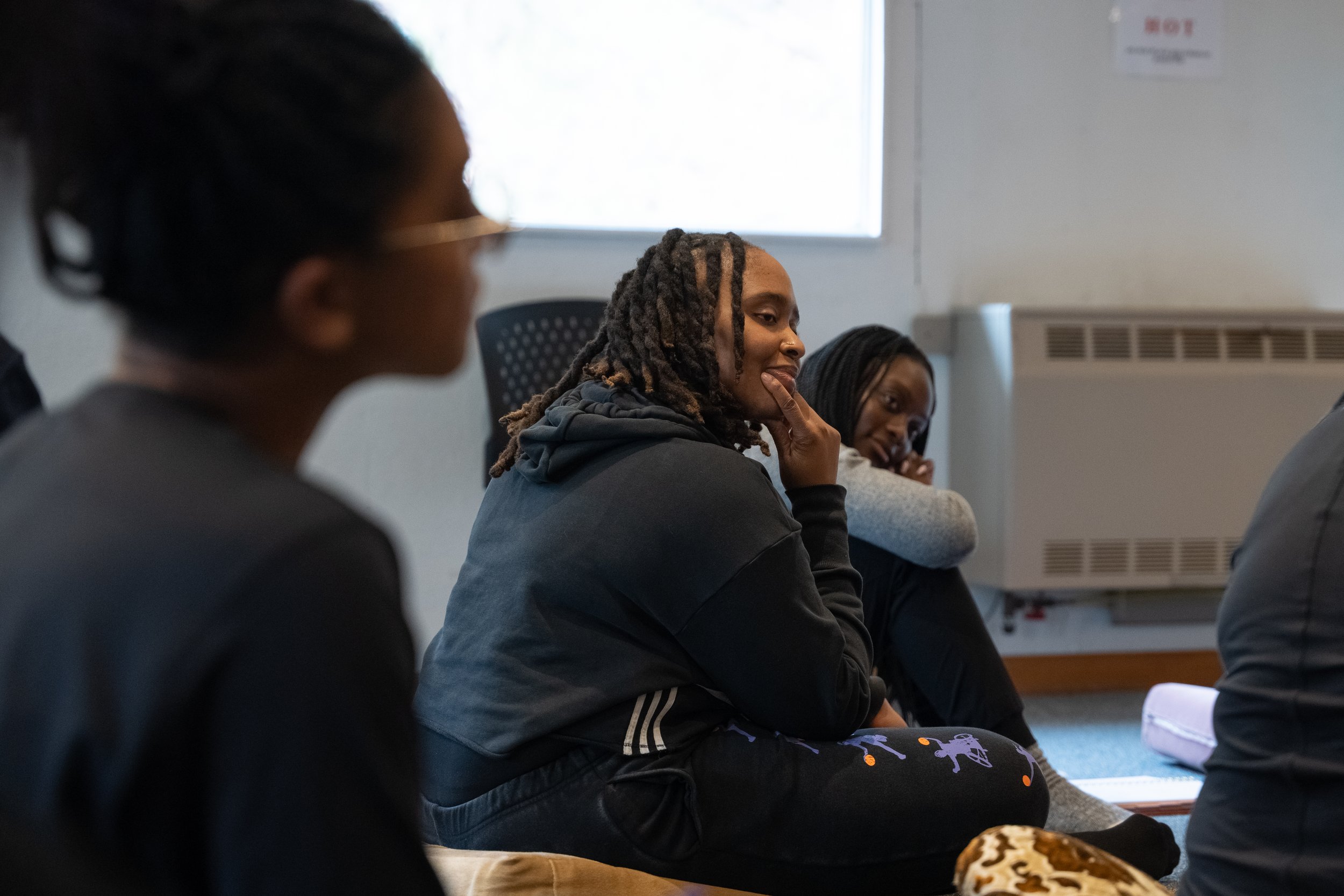 A group of women sitting on the floor attending a discussion or class, with a woman in the middle smiling and resting her chin on her hand.