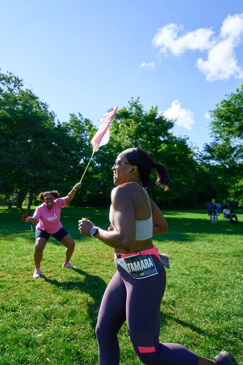 Two women participating in a race or running event in a park on a sunny day, with one woman running in the foreground wearing a bib that says 'TAMARA' and the other woman in the background waving a pink and white flag, both engaged and happy.