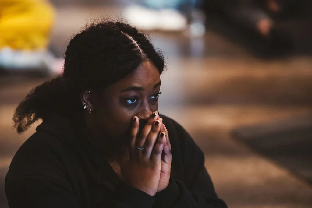 A woman with dark hair in braids, wearing earrings, covering her mouth with her hands, with a worried or emotional expression, in a dimly lit indoor setting.