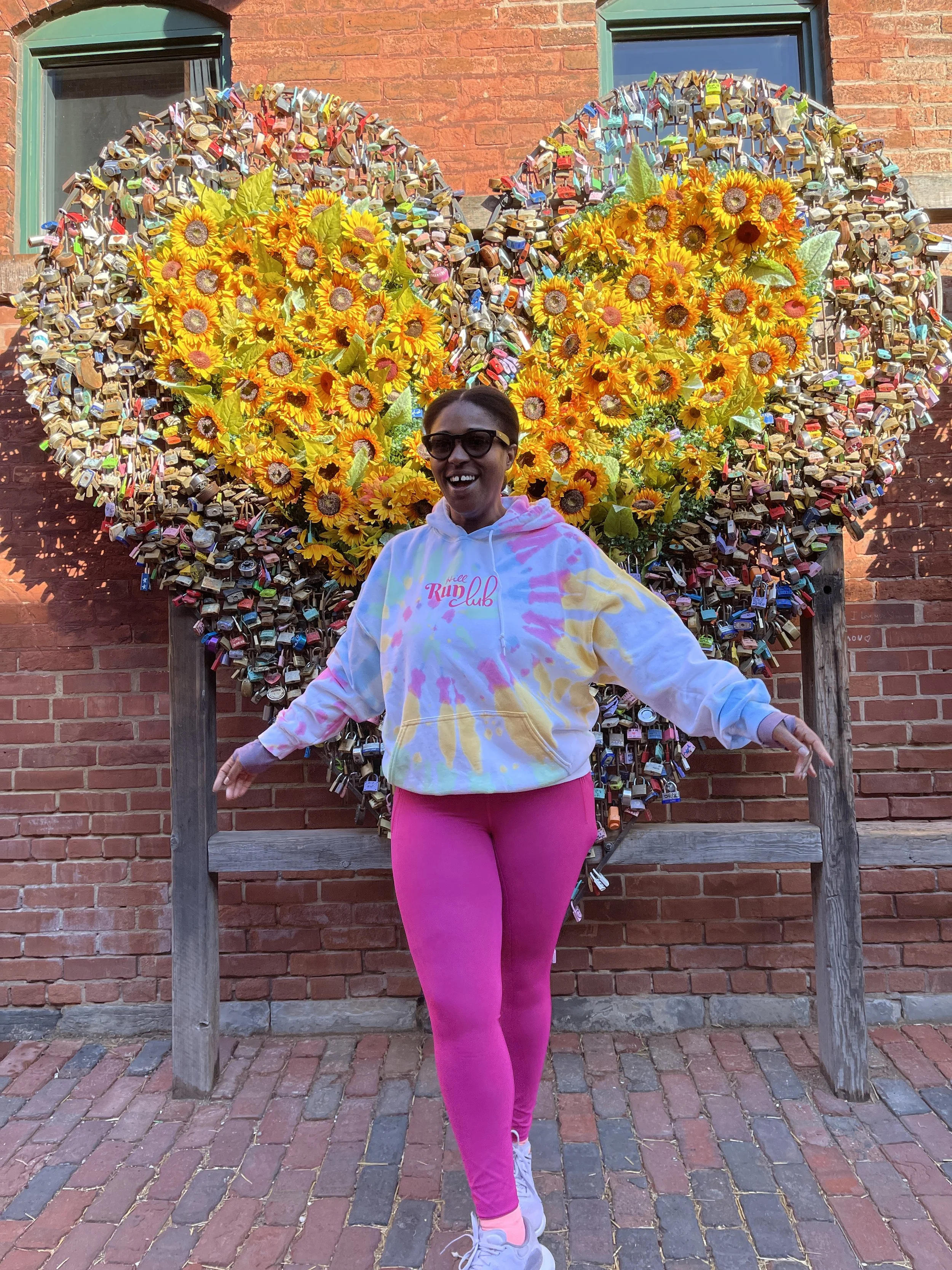 A woman wearing sunglasses, a tie-dye hoodie, and pink leggings smiling with arms outstretched in front of a large decorative heart made of yellow sunflowers and numerous padlocks on a brick wall.