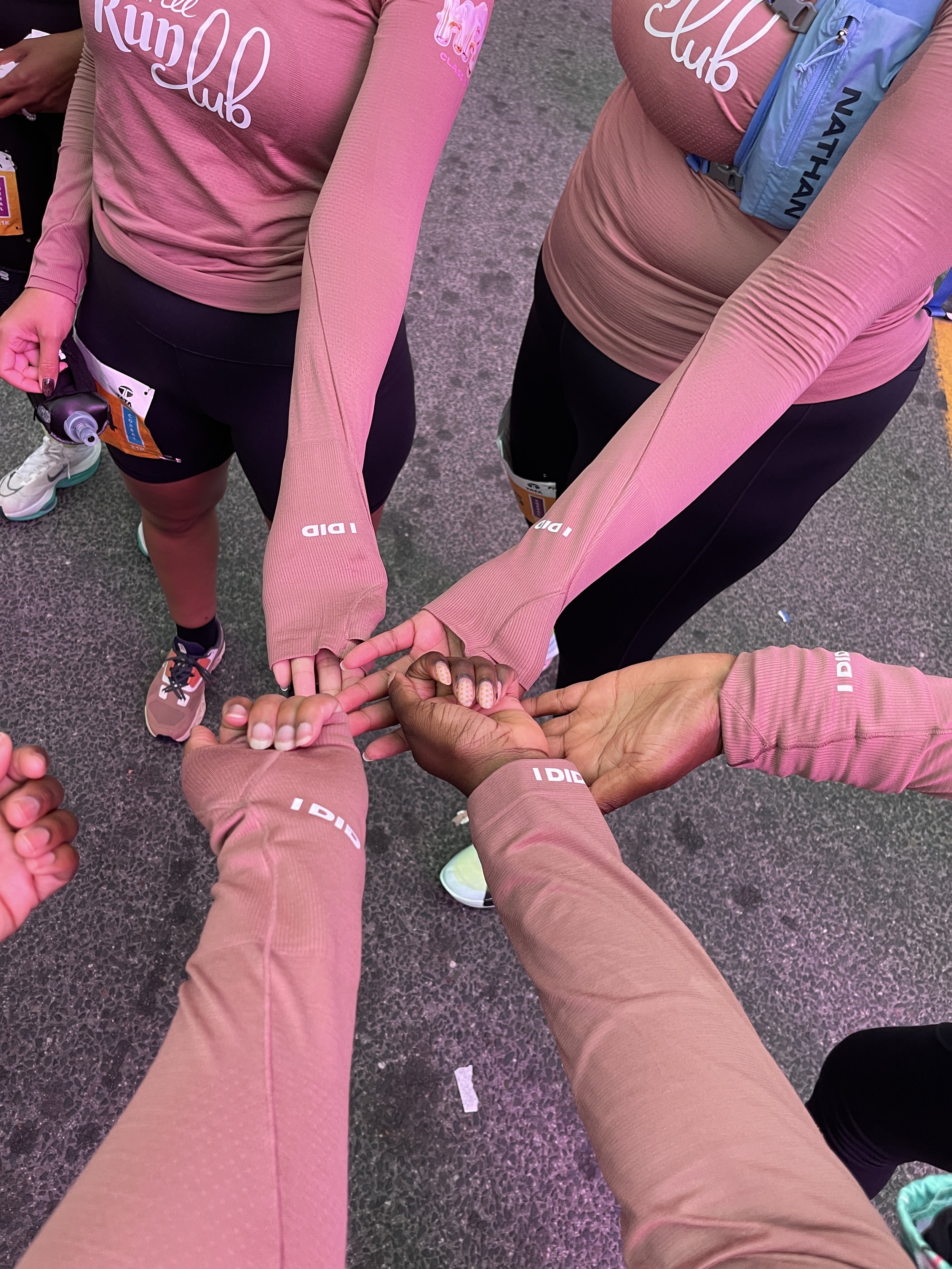 Group of four people wearing matching pink athletic long-sleeve shirts and black shorts, joining hands in a pile.