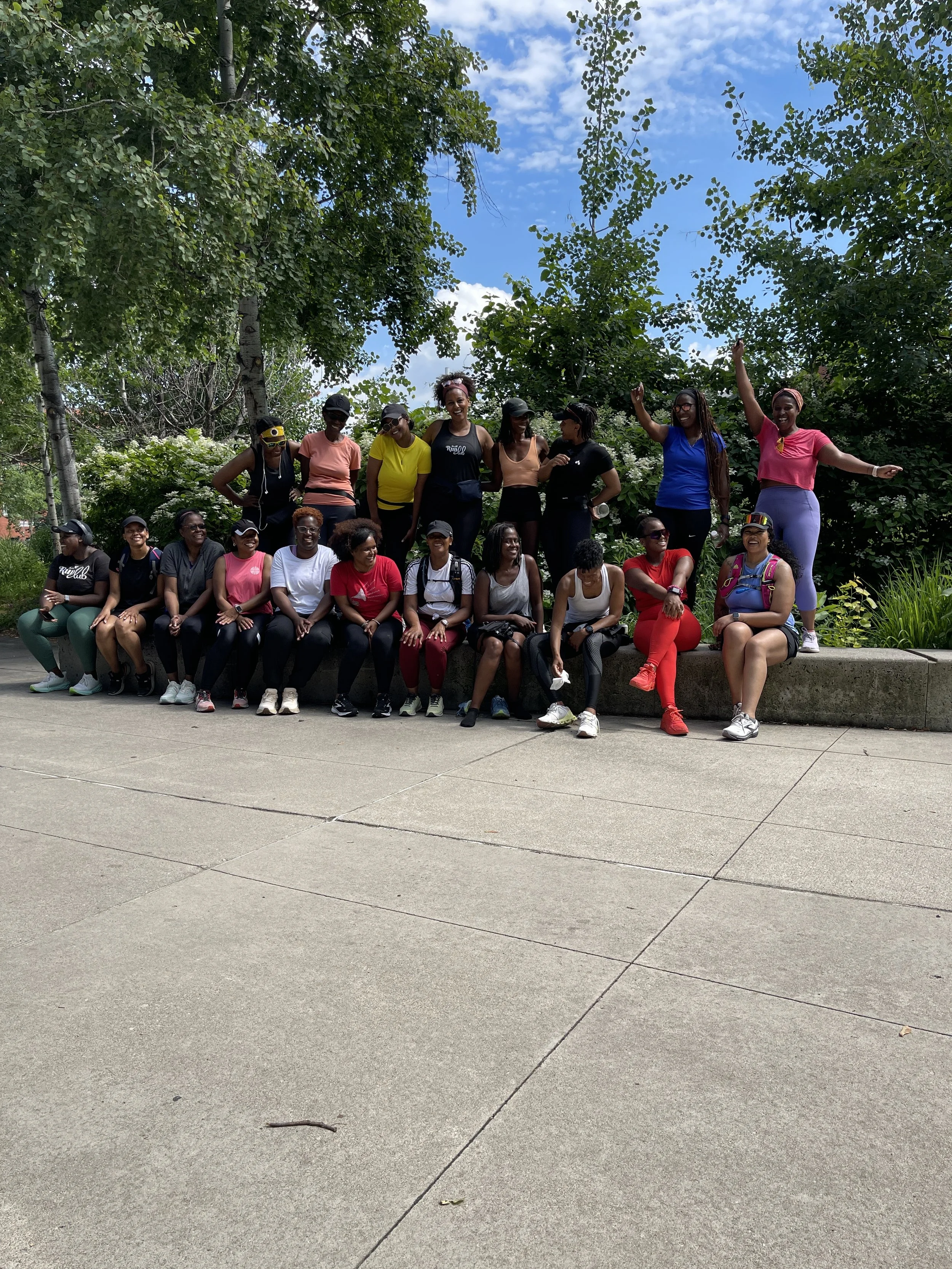 Group of women in athletic clothing sitting and standing on a ledge outdoors, some waving or smiling, with trees and a blue sky with clouds in the background.