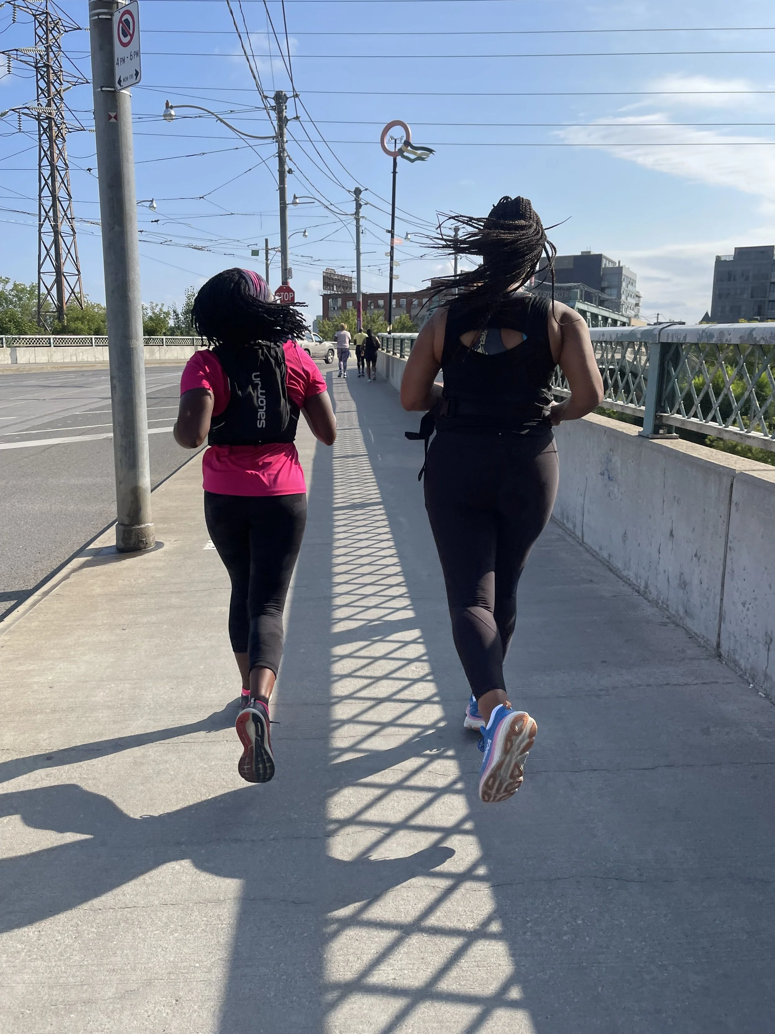 Two women jogging on a city sidewalk under a clear blue sky with shadows cast by a metal railing and utility poles.