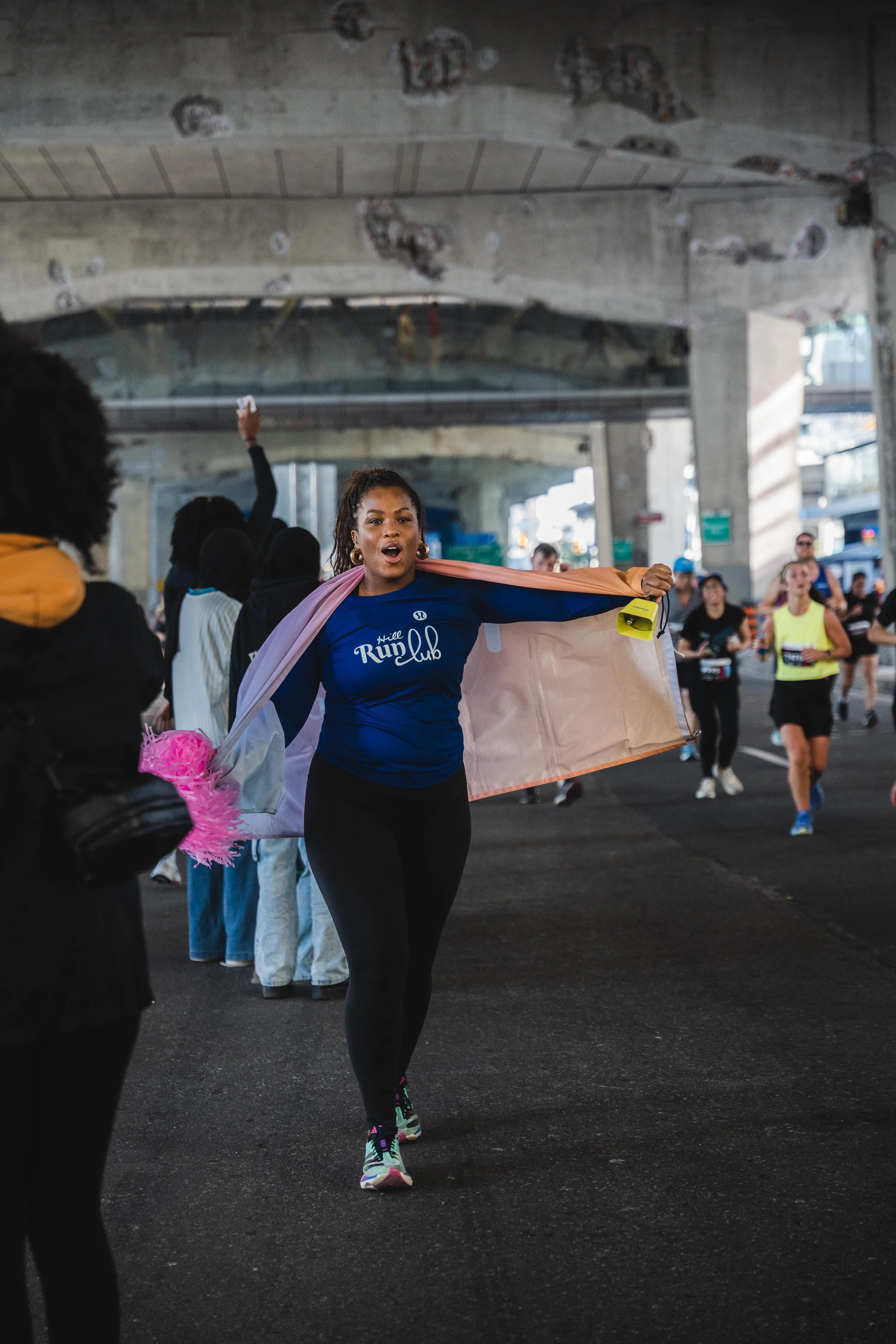 A woman at a running event holding a flag with a group of runners in the background under a bridge.