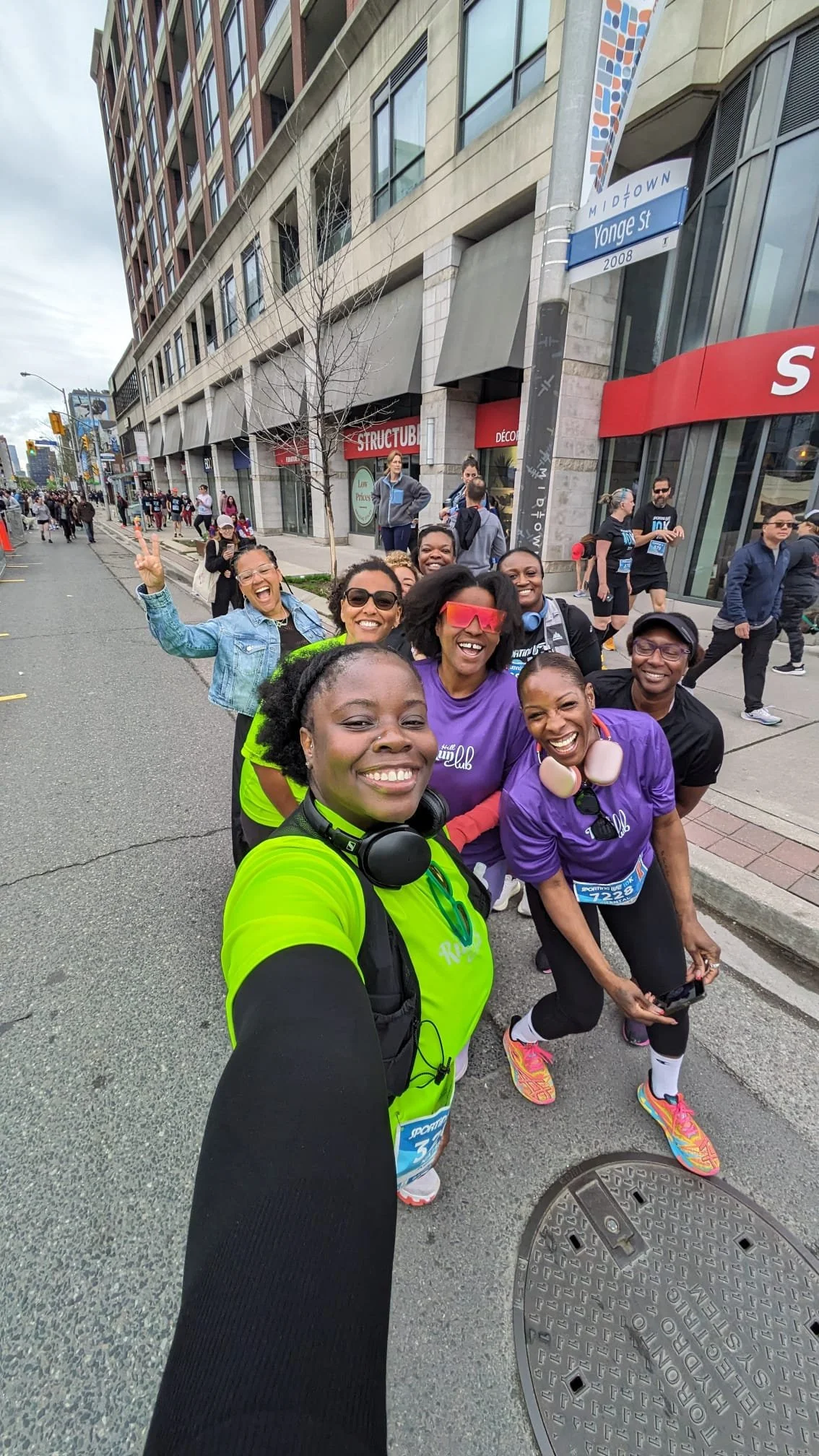 Group of smiling women taking a selfie on a city street during a marathon or running event, with some wearing colorful running attire and headphones.