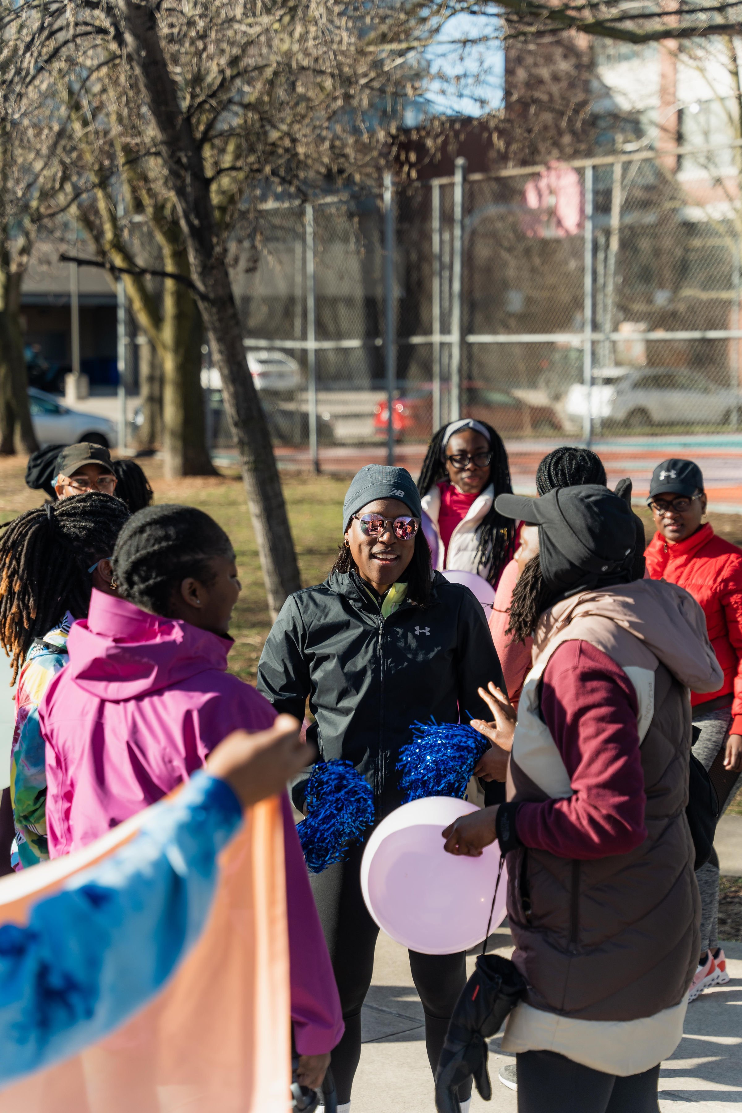 A group of women gathered outdoors in a park, wearing athletic clothing and holding balloons, engaging in conversations on a sunny day near a wooded area and a fenced tennis court.