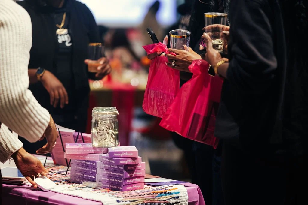 People at a table with pink gift bags and books, holding glasses with drinks, at a social event.