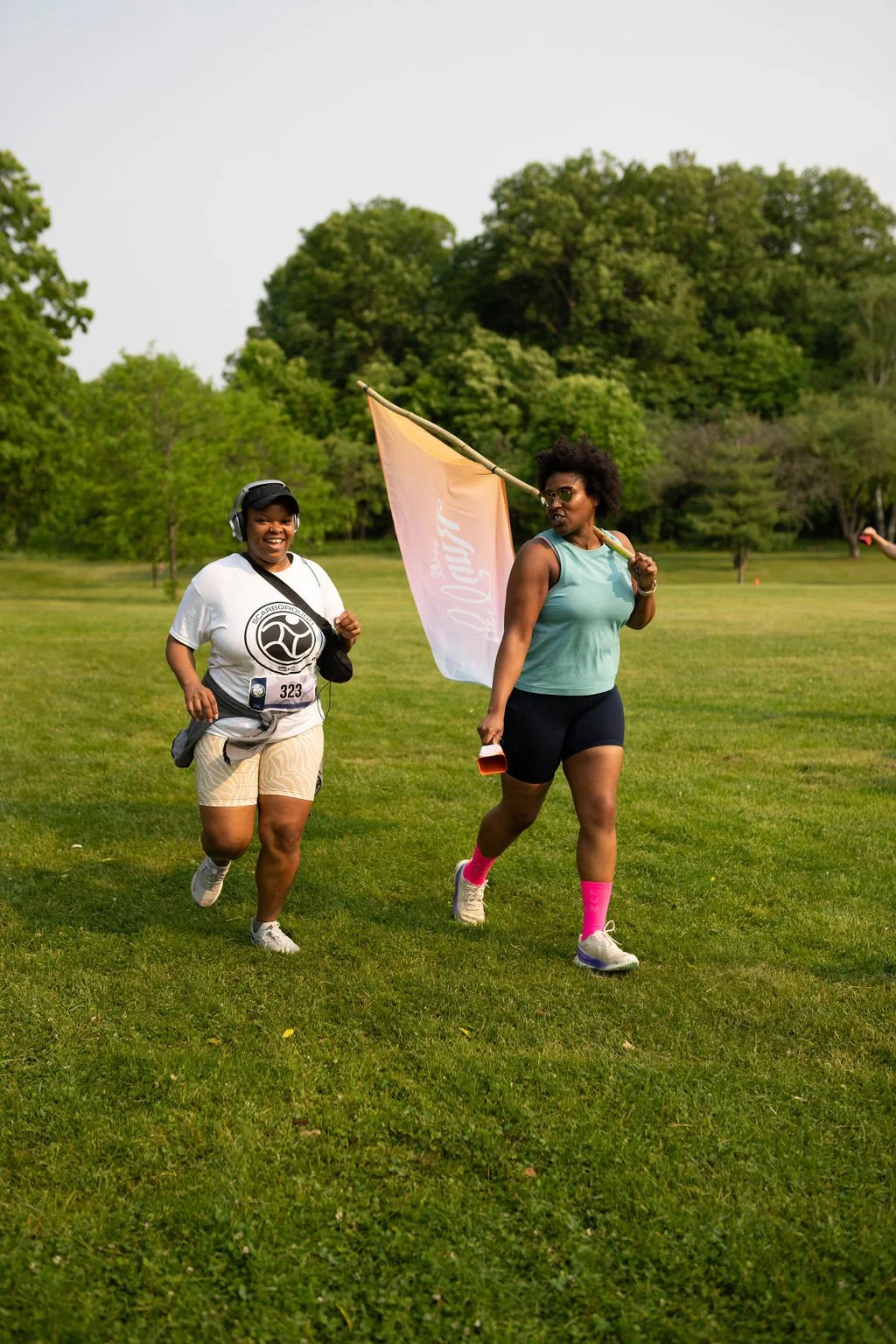 Two women running outdoors on a grassy field, one holding a flag, with trees in the background during daytime.