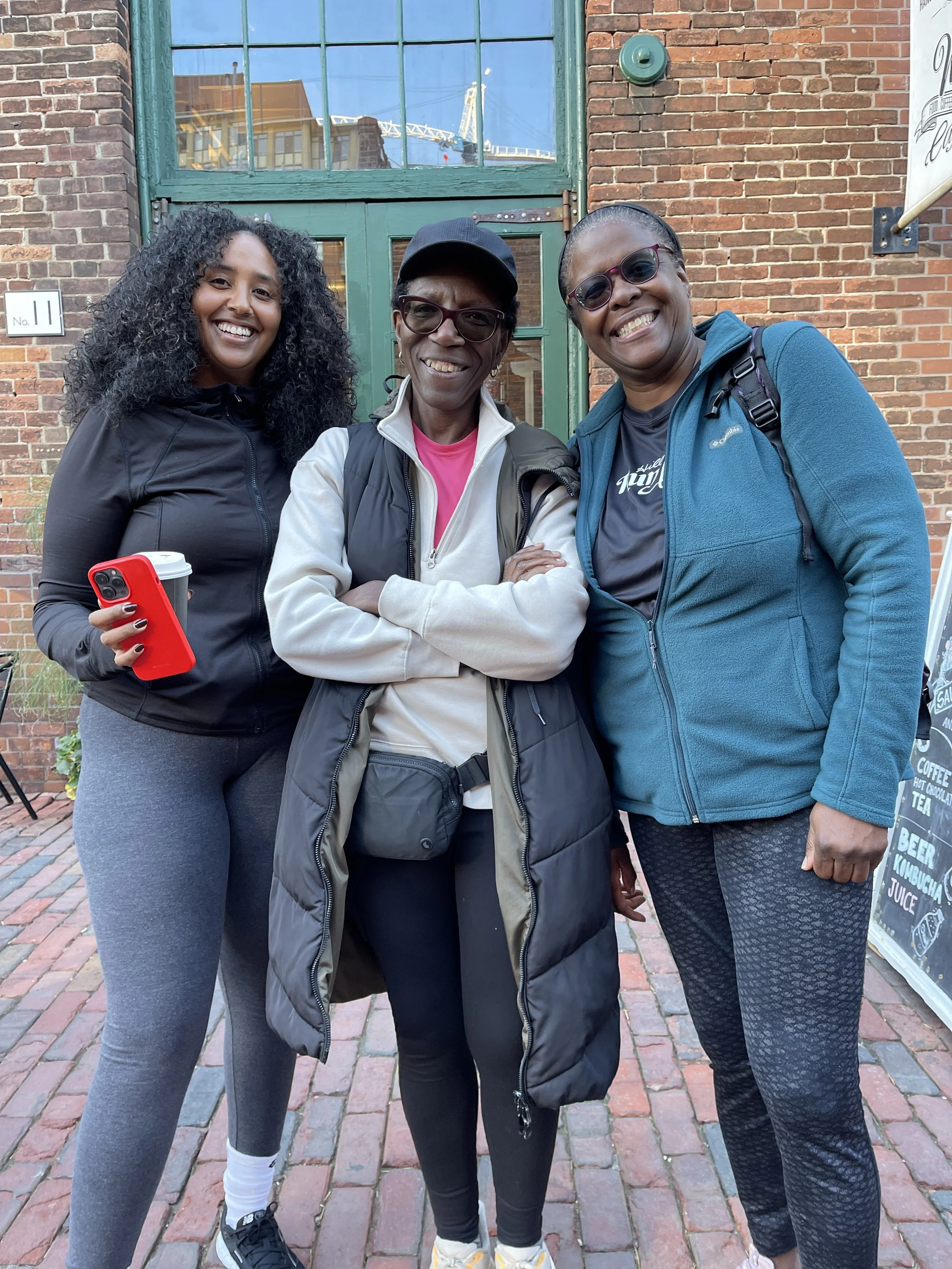 Three women standing outdoors in front of a brick building, smiling, with one woman holding a red phone and coffee cup.