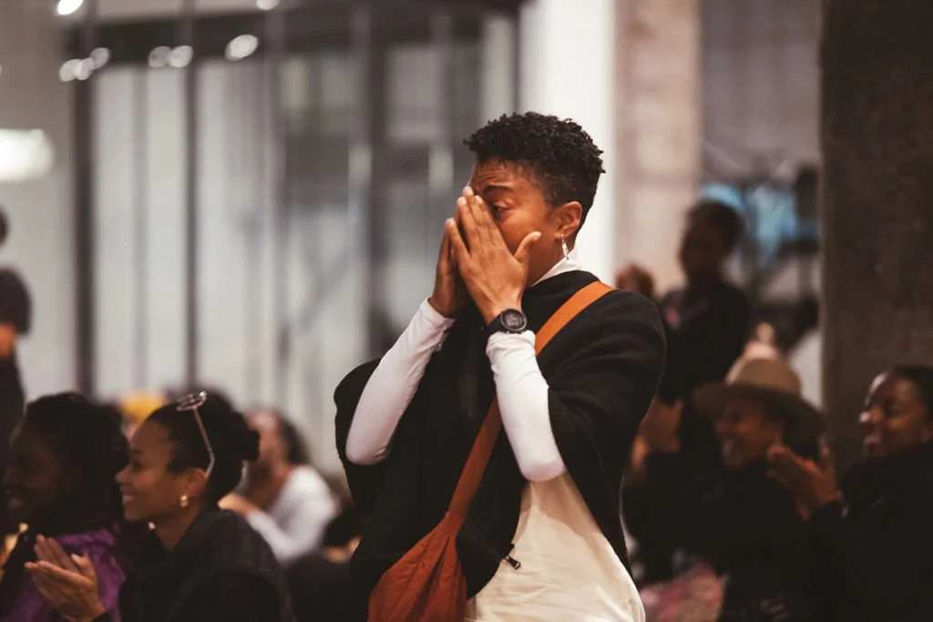 A woman with short curly hair covering her face with her hands, standing in a busy indoor setting with seated and standing people around her, some smiling and clapping, in a social or public gathering.