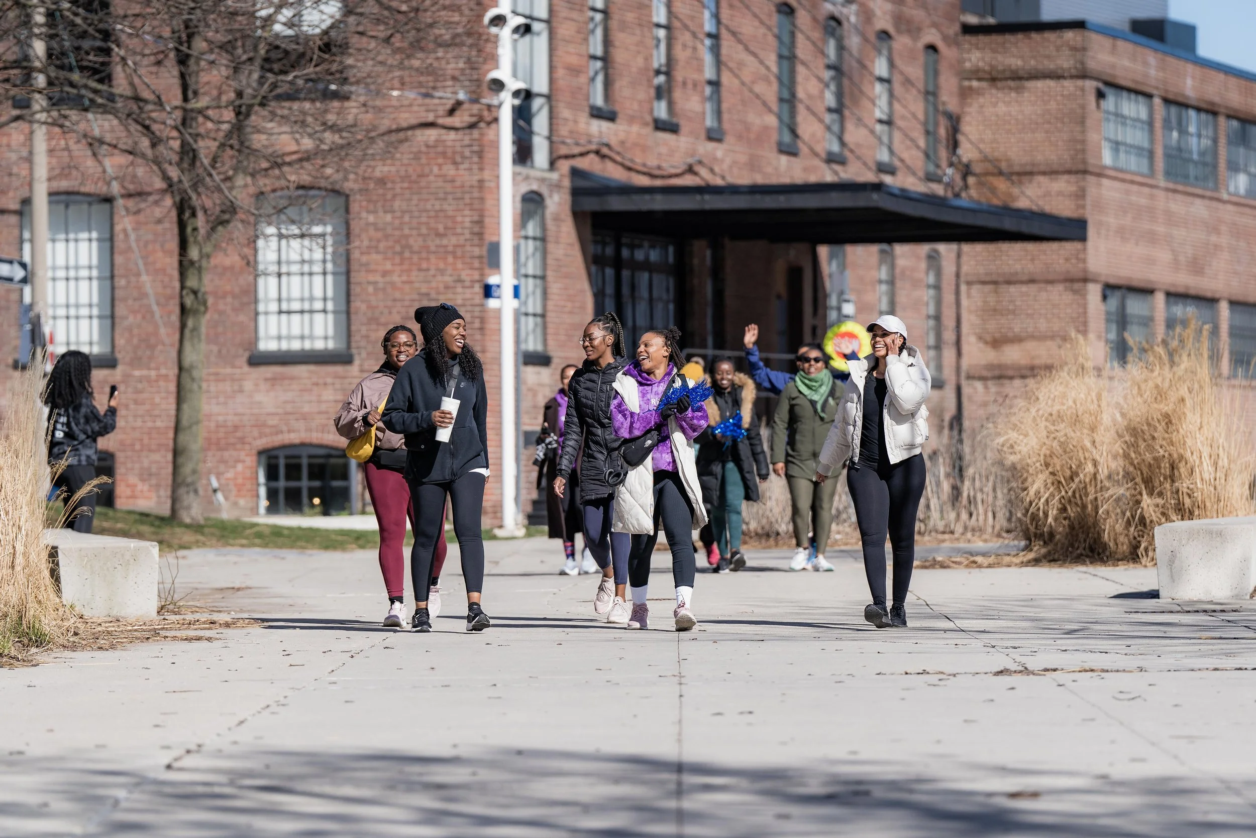 Group of diverse women walking and smiling outdoors in front of a brick building on a sunny day.
