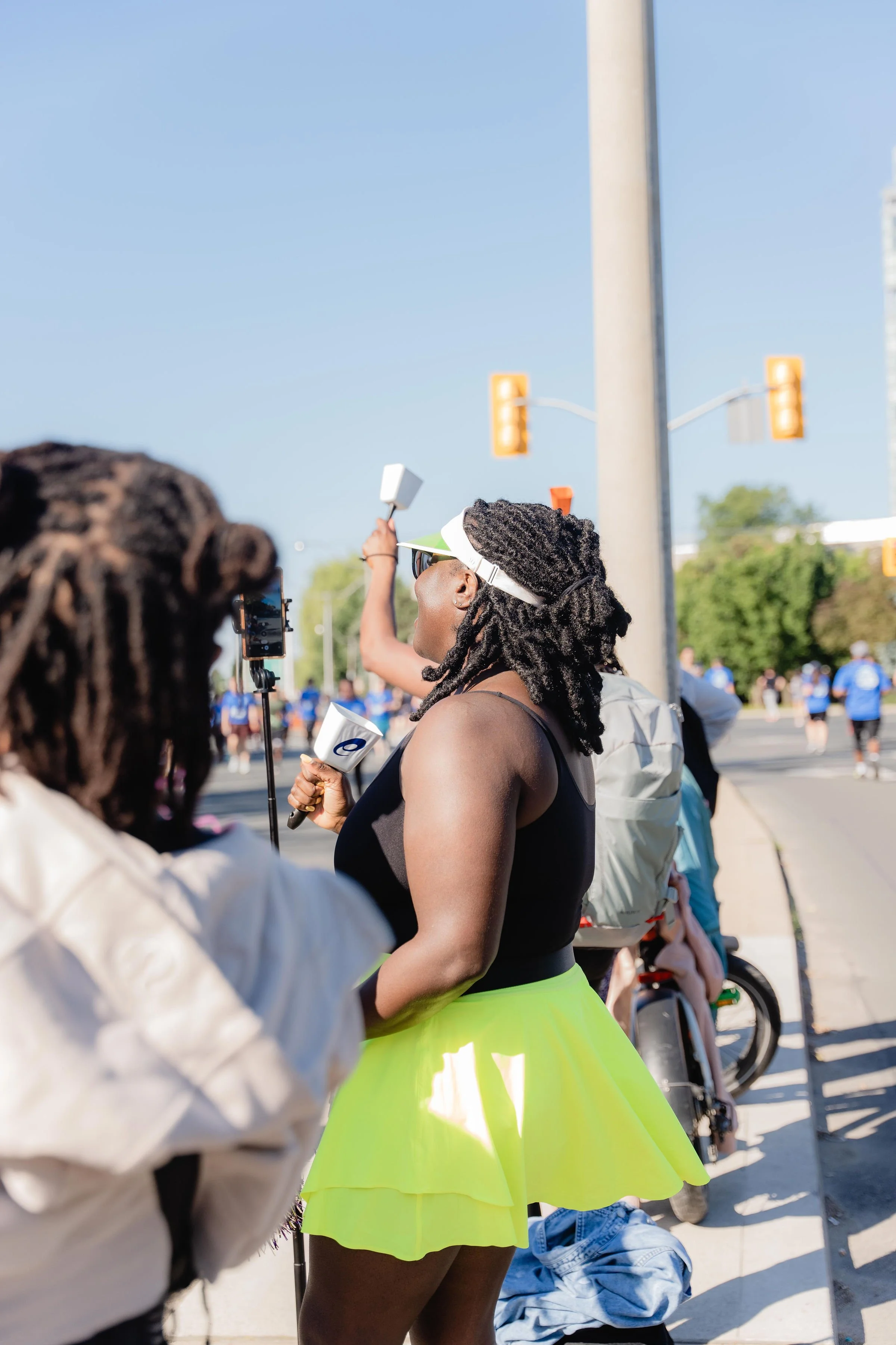 A woman with dreadlocks wearing a black top and neon yellow skirt stands on a sidewalk at a public event, holding a cup and a small flag, with a crowd of people and traffic lights in the background.