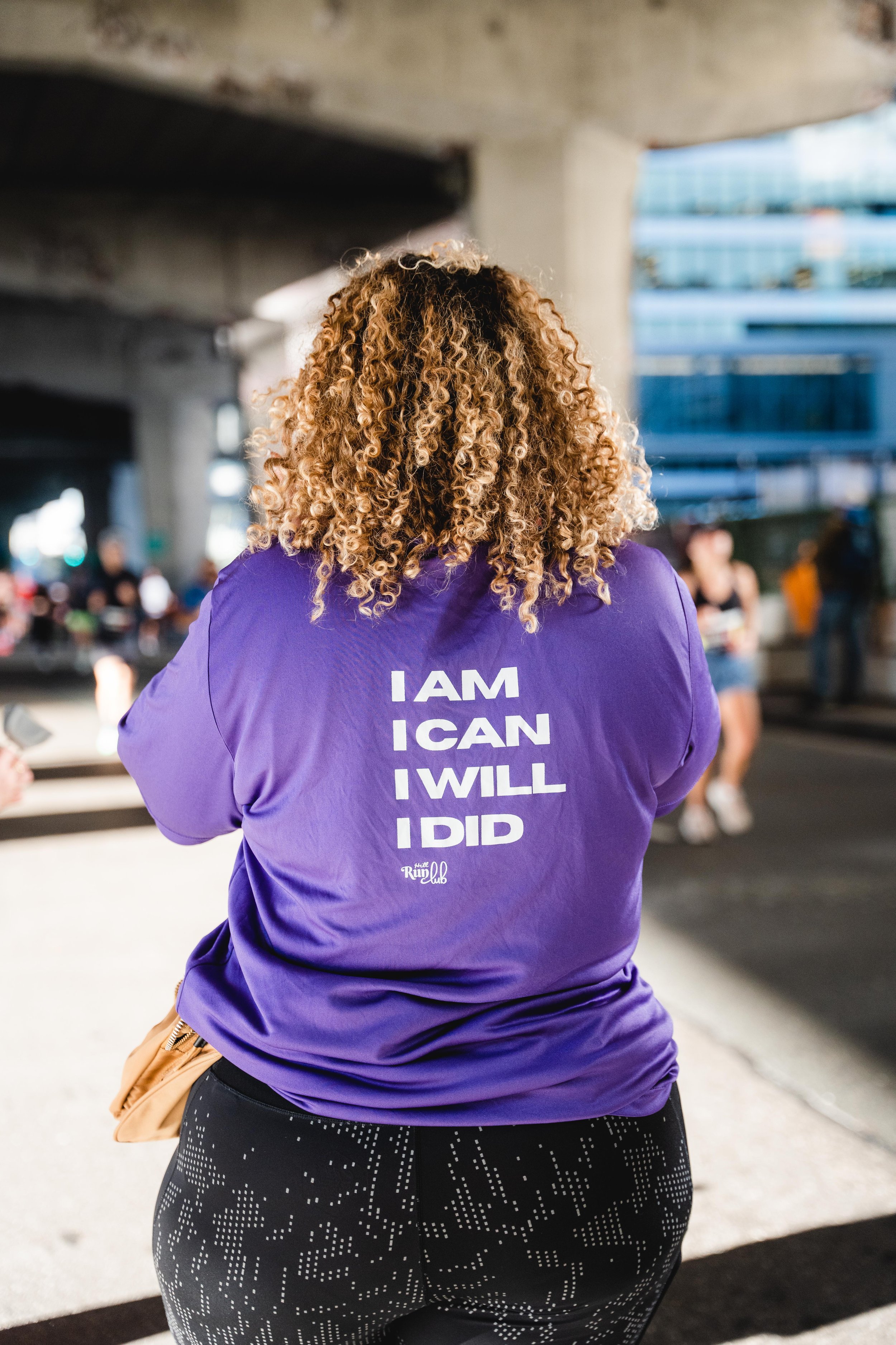 Back view of a woman wearing a purple shirt with an inspirational quote and black leggings with white dotted patterns, standing under a bridge or overpass.
