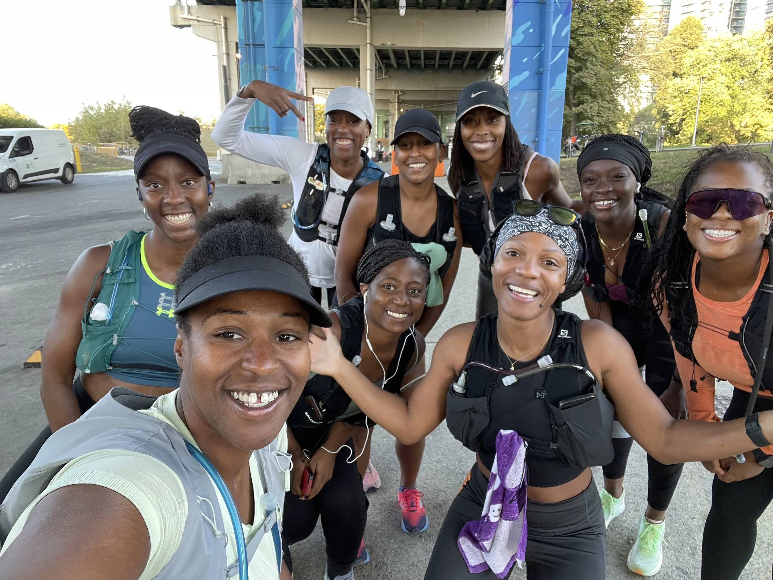 A group of ten diverse women dressed in athletic gear, smiling and posing for a selfie outdoors under a bridge, with trees and a partly cloudy sky in the background.