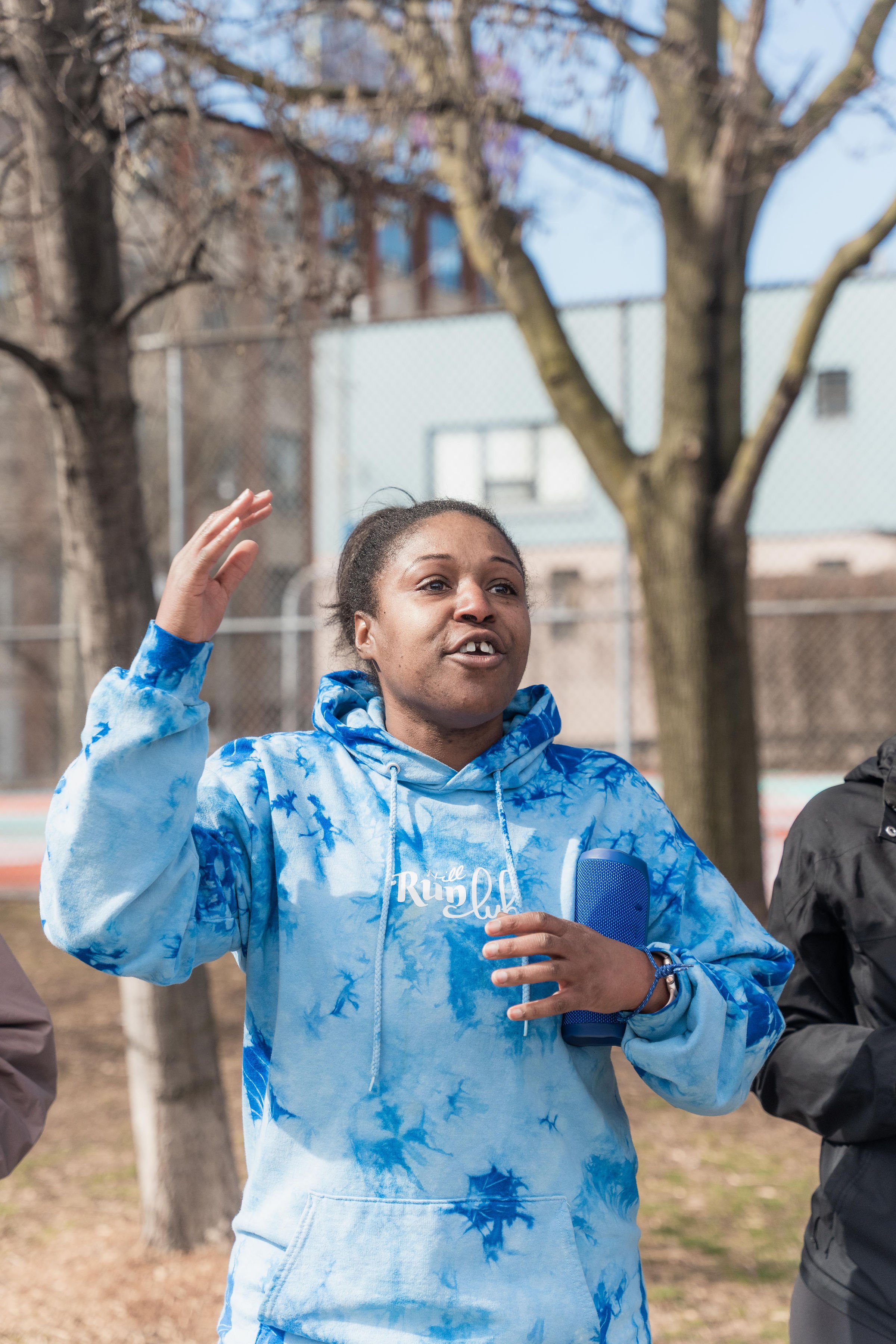 A woman in a blue tie-dye hoodie with 'Run Club' written on it, speaking outdoors in a park with trees and a chain-link fence.