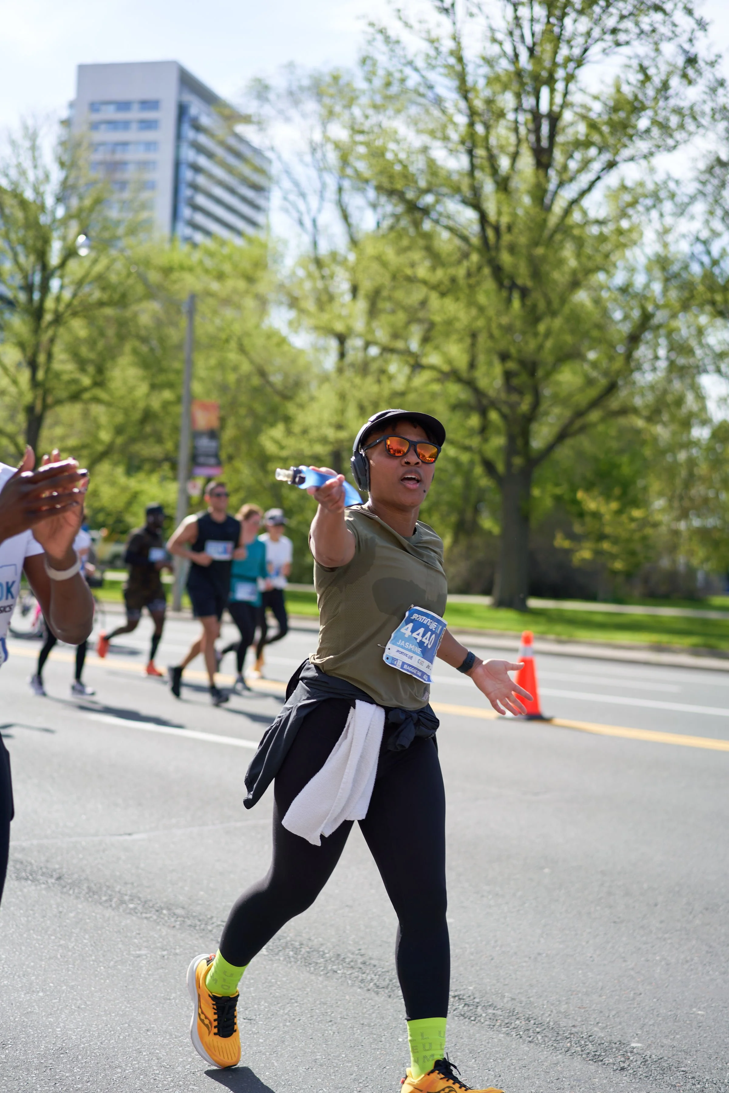 A woman running a marathon on a city street during daytime, pointing at the camera, wearing sunglasses, headphones, and a race bib.