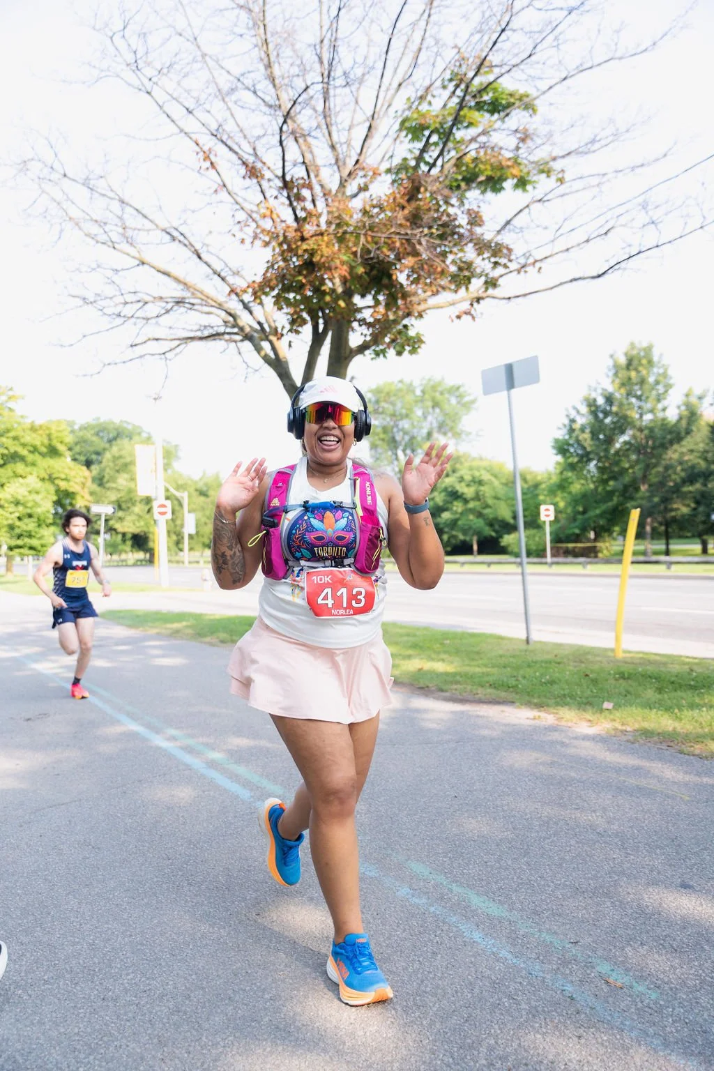 A woman running in a 10K race, wearing a white tank top, light pink skirt, and blue running shoes, with a pink hydration pack, sunglasses, and a white cap, smiling and waving during the race in a park with trees and a blue sky.