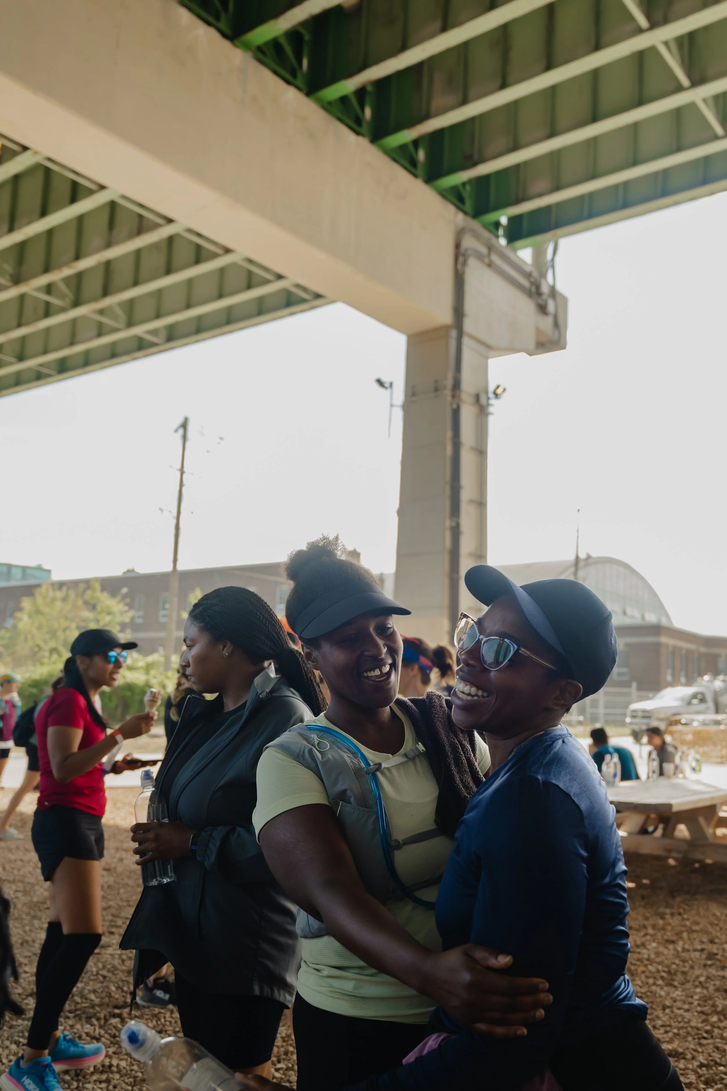 Two women smiling and hugging under a bridge during an outdoor event, with other people in the background.