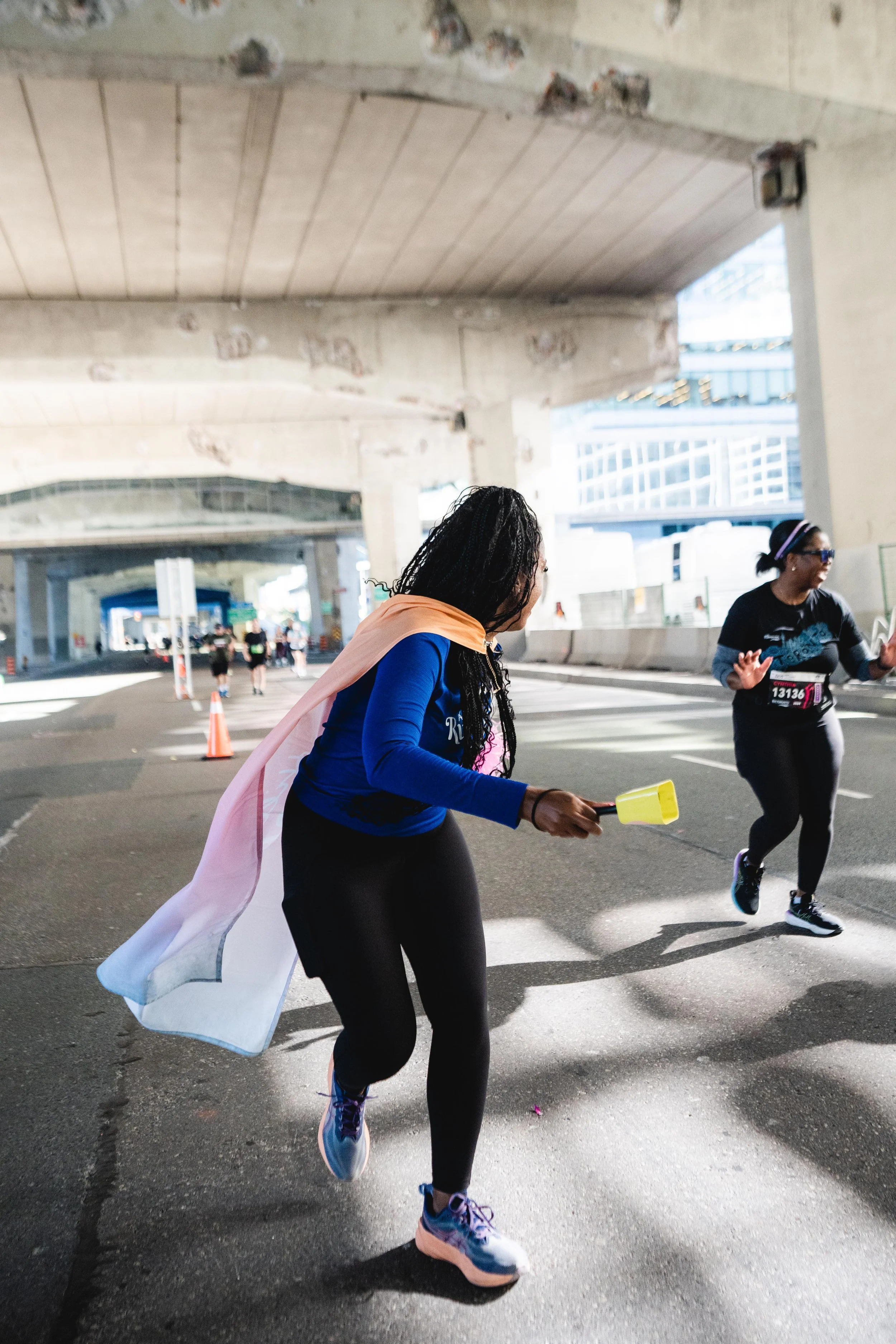 A woman wearing black leggings, a blue long-sleeve shirt, and a cape running in a marathon or race under an overpass. She is holding a yellow object in her hand. Other runners are visible in the background.