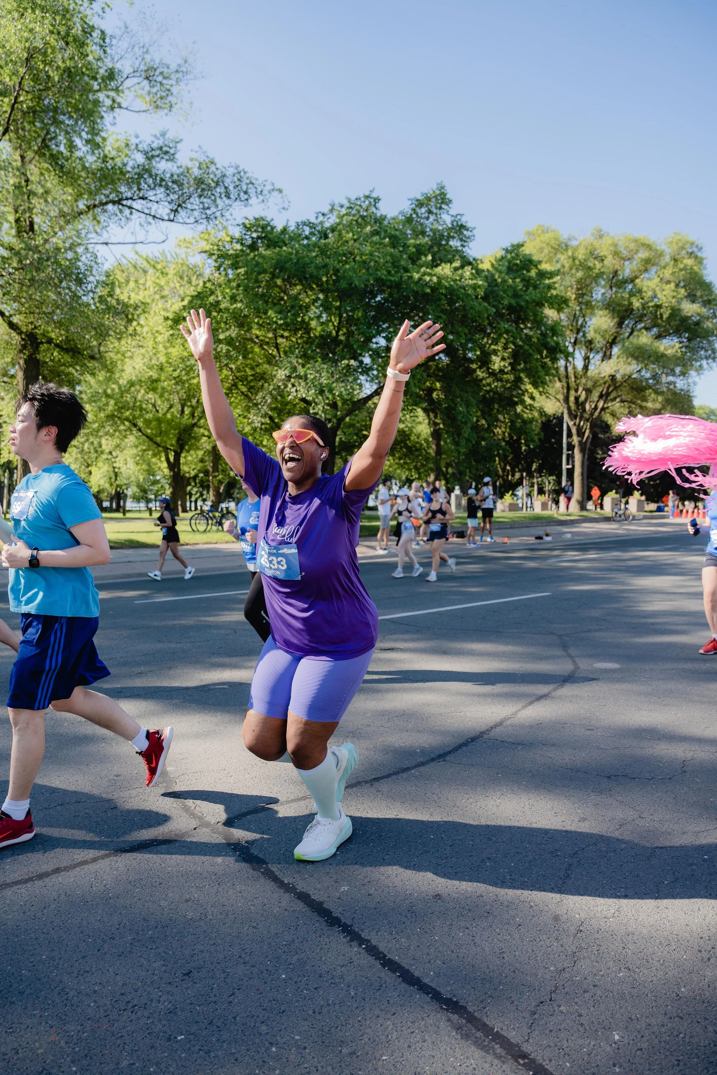 A smiling woman in a purple shirt and shorts participating in a marathon, raising her arms in excitement, surrounded by other runners in a park with green trees on a sunny day.