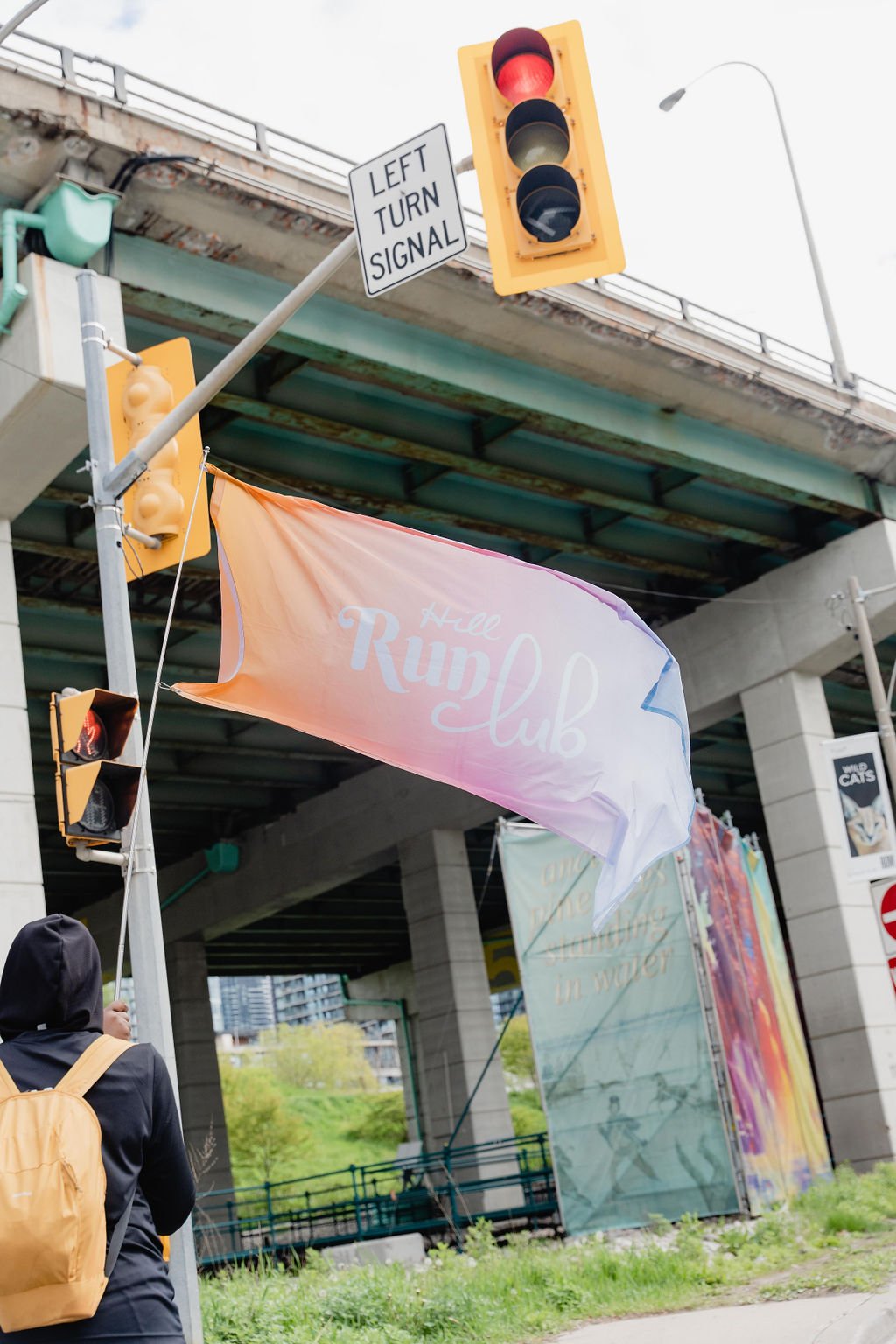 Urban street scene featuring traffic lights, a sign for a left turn signal, a colorful flag that reads 'Hill Run Club', and an overpass. A person with a backpack is visible from behind.