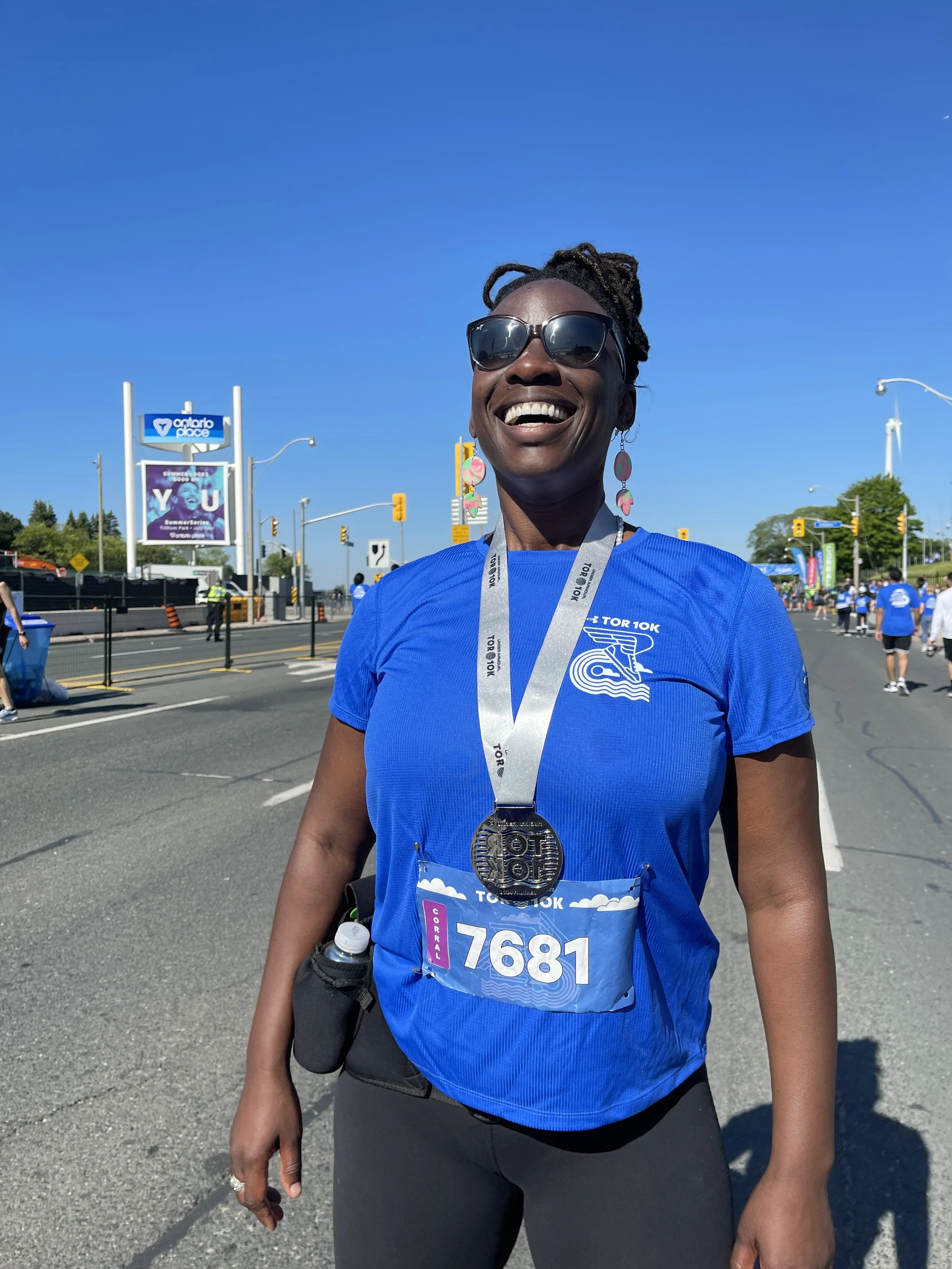 A smiling woman wearing sunglasses, a blue race shirt, and a bib number 7681 with a medal around her neck standing on a city street during a running event.