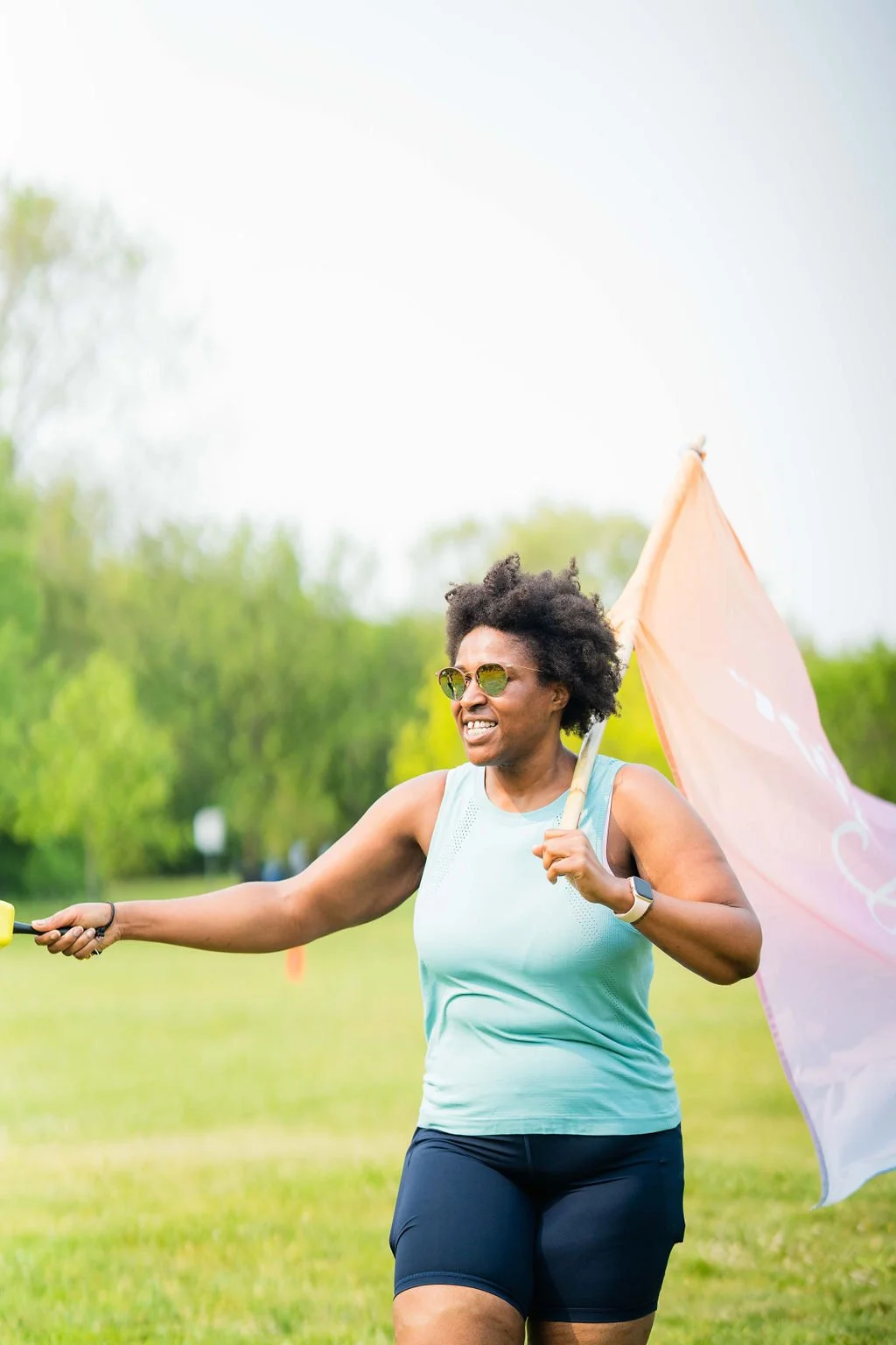 A woman with short curly hair, sunglasses, and athletic clothing is running outdoors while holding a pink flag, smiling, in a grassy park with green trees in the background.