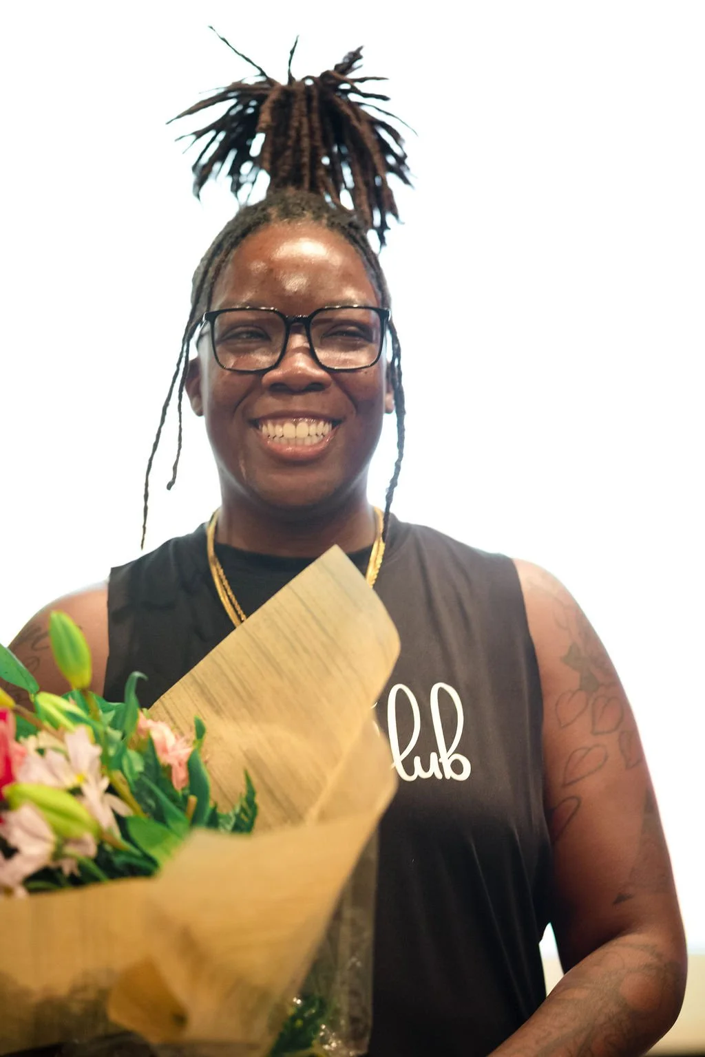 A woman smiling while holding a bouquet of flowers, wearing glasses and a sleeveless black shirt, with tattoos on her arm and a large hairstyle that looks like dreadlocks tied up at the top.