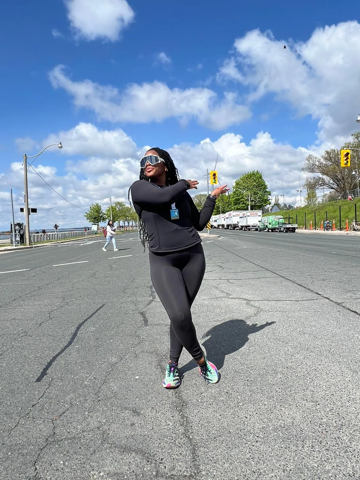 A woman in black athletic wear and sunglasses standing on an empty city street with traffic lights, trees, and a blue sky with clouds.