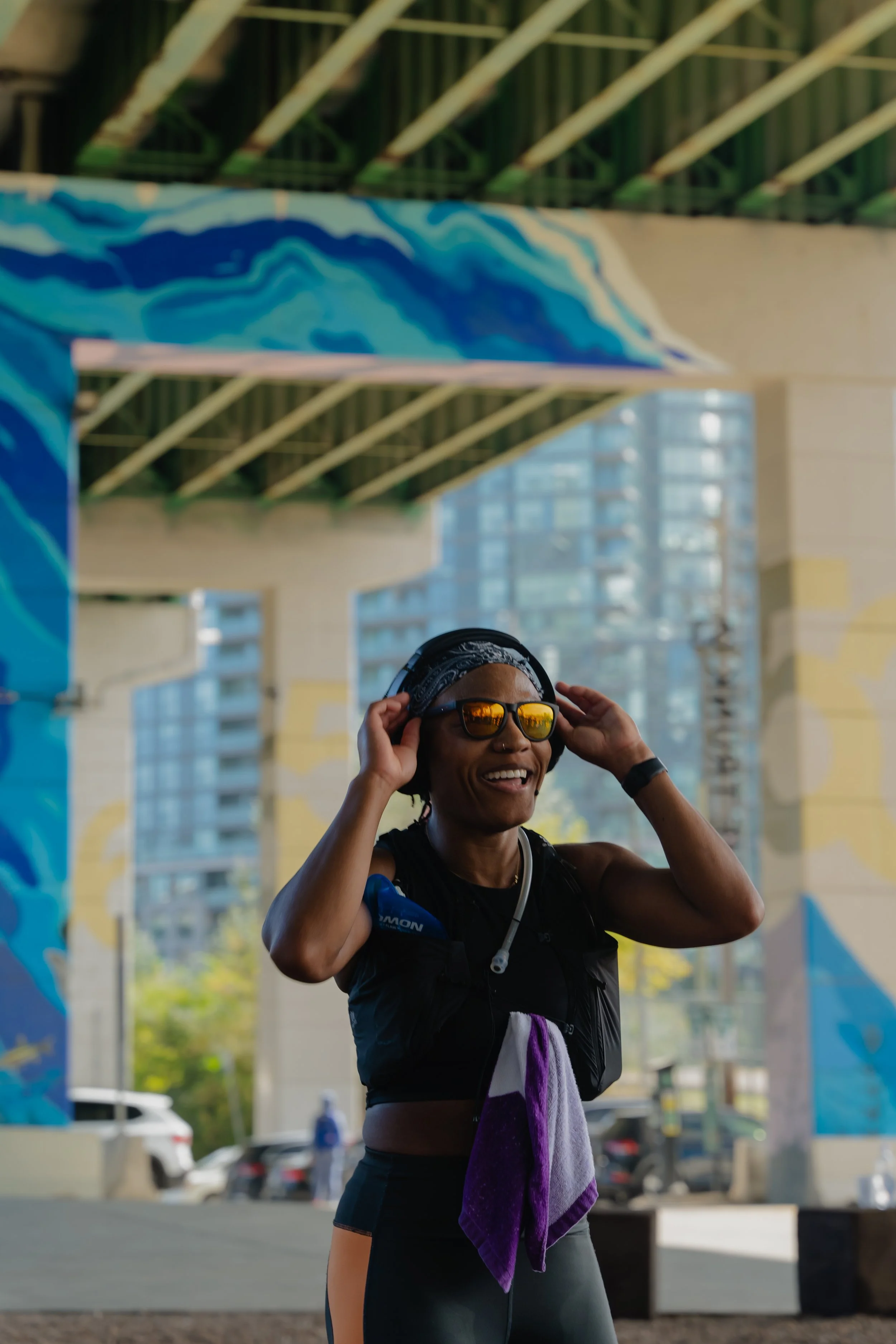 Woman wearing sunglasses and headphones smiling, standing under a colorful mural, holding her headphones with both hands.