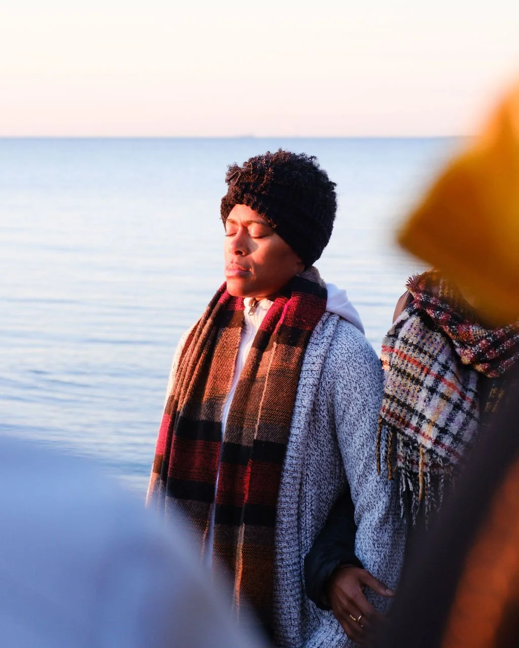 A woman standing by a body of water with her eyes closed, wearing a black knit hat, a plaid scarf, and a gray coat.