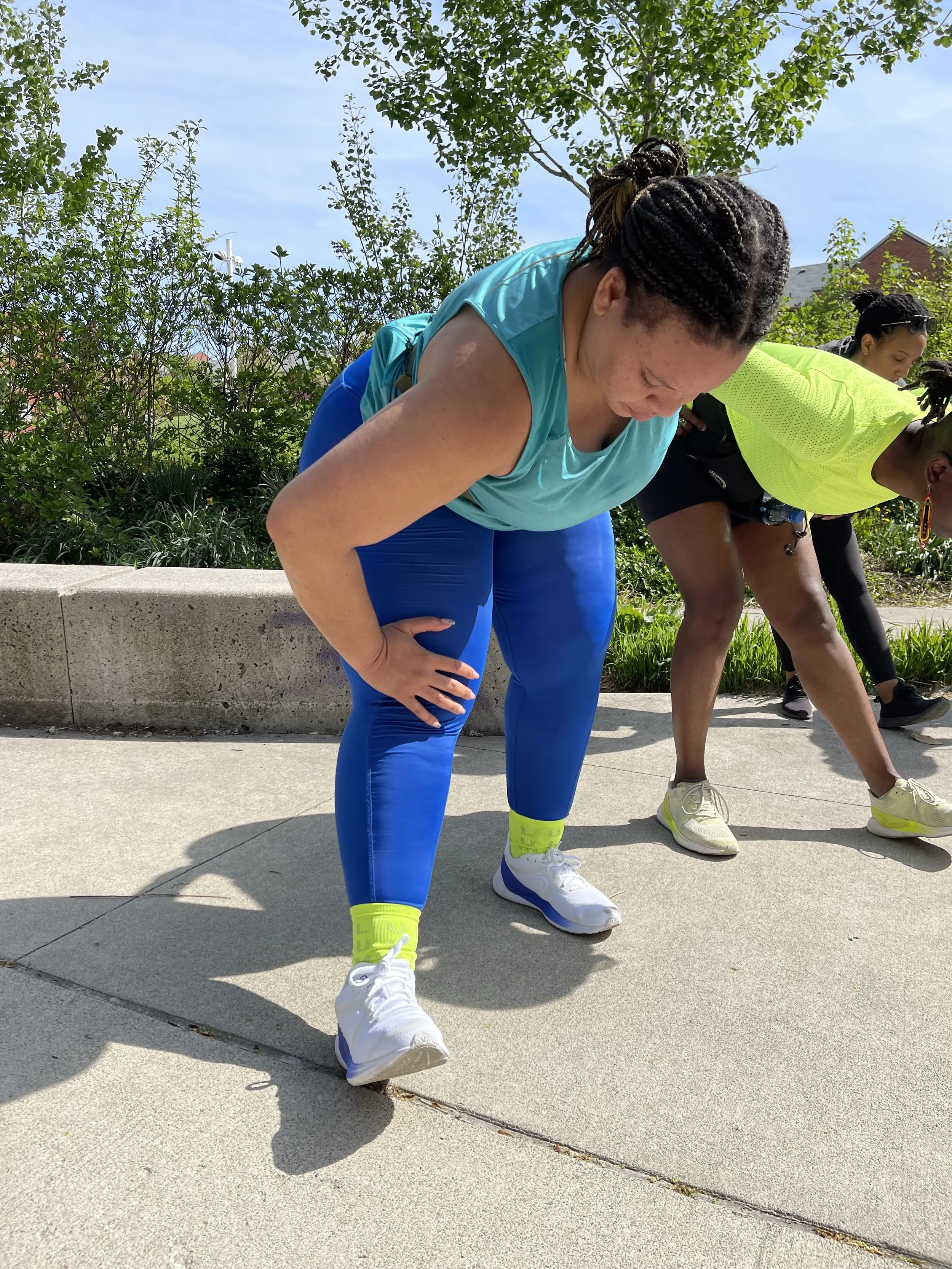Hill Run Club warm up. Women stretching outdoors on a sunny day, with one woman in a turquoise shirt and blue leggings in the foreground, and others in the background, all bending forward with one hand on their knees.