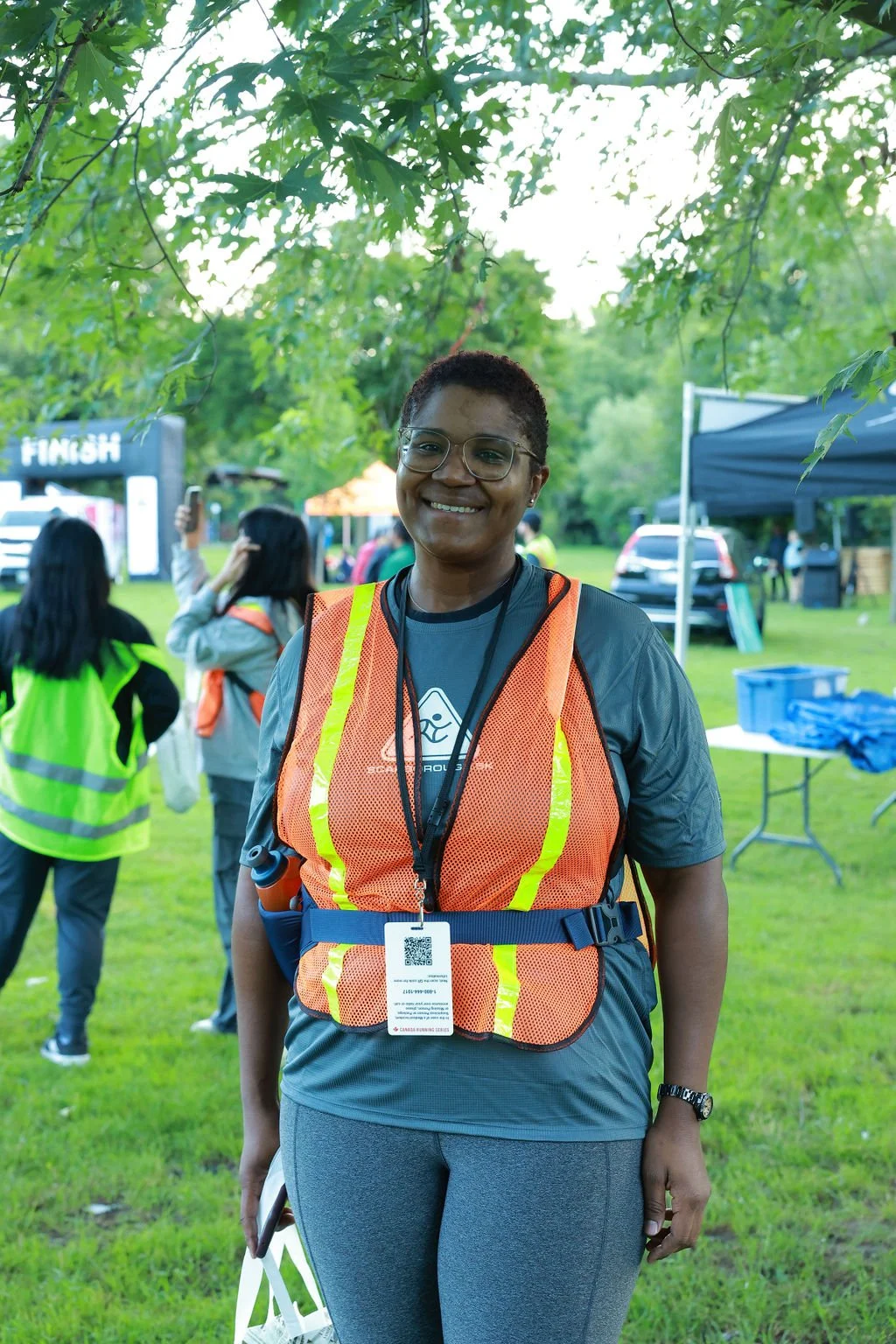 Smiling woman wearing glasses and a reflective safety vest at an outdoor event near a finish line, with several people and tents in the background.
