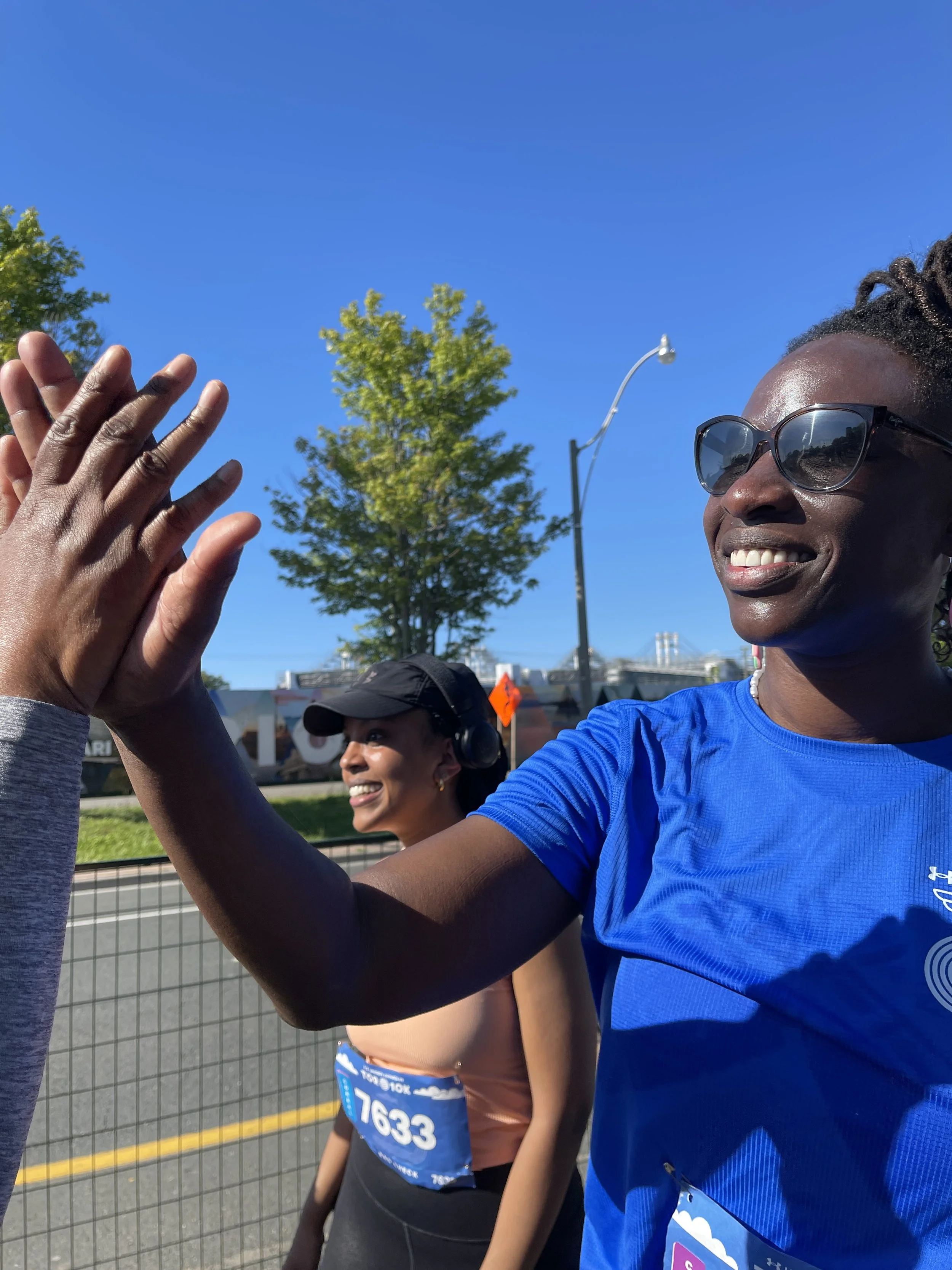 Two women high-fiving each other during a race or marathon, with race bibs and a smiling woman in the background, outdoors on a sunny day.