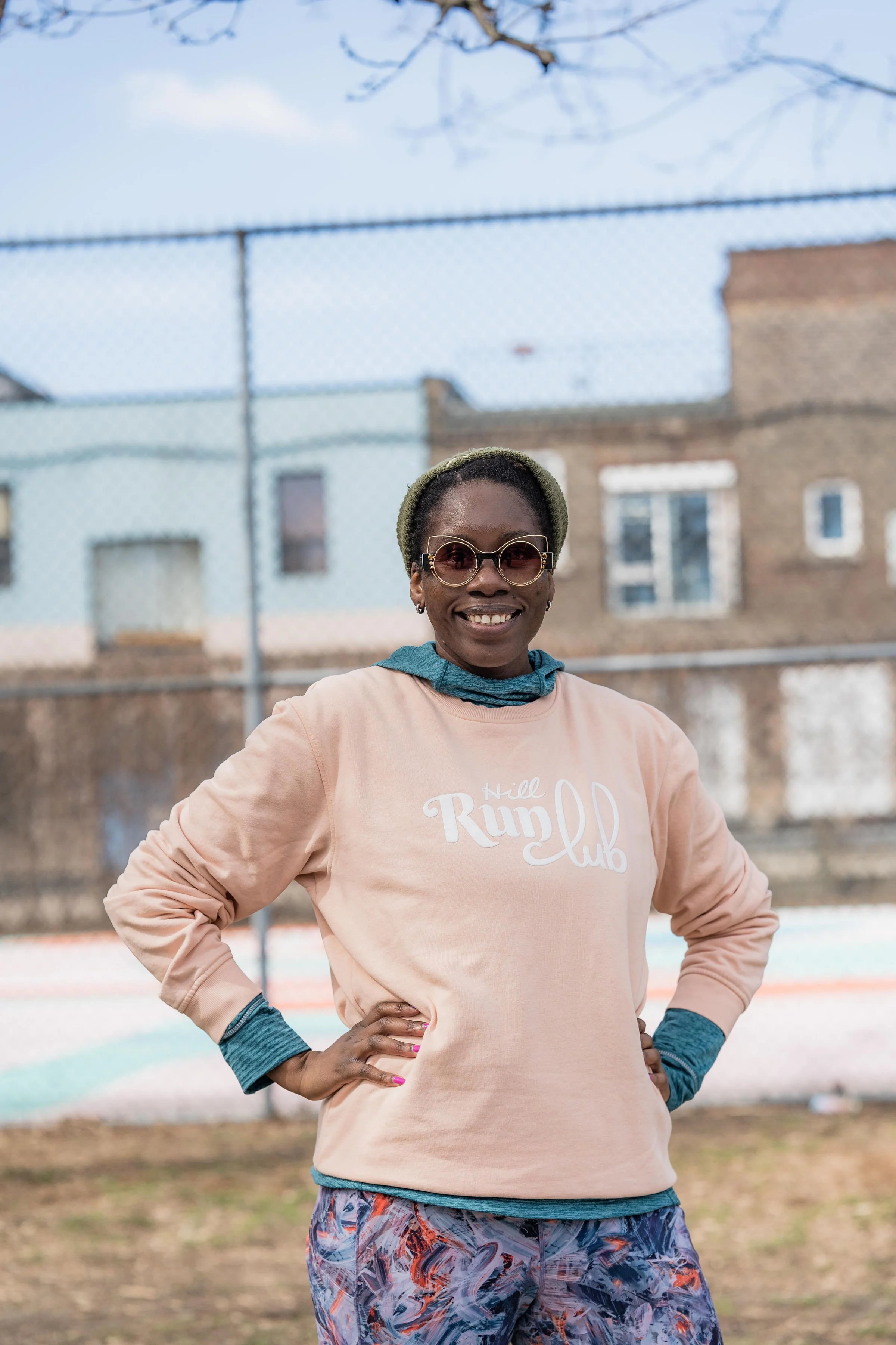 A smiling woman standing with hands on hips outdoors, wearing sunglasses, a beige sweatshirt with 'Hill Run Club' written on it, colorful shorts, and a headband, in front of a chain-link fence with a brick building and a cloudy sky in the background.