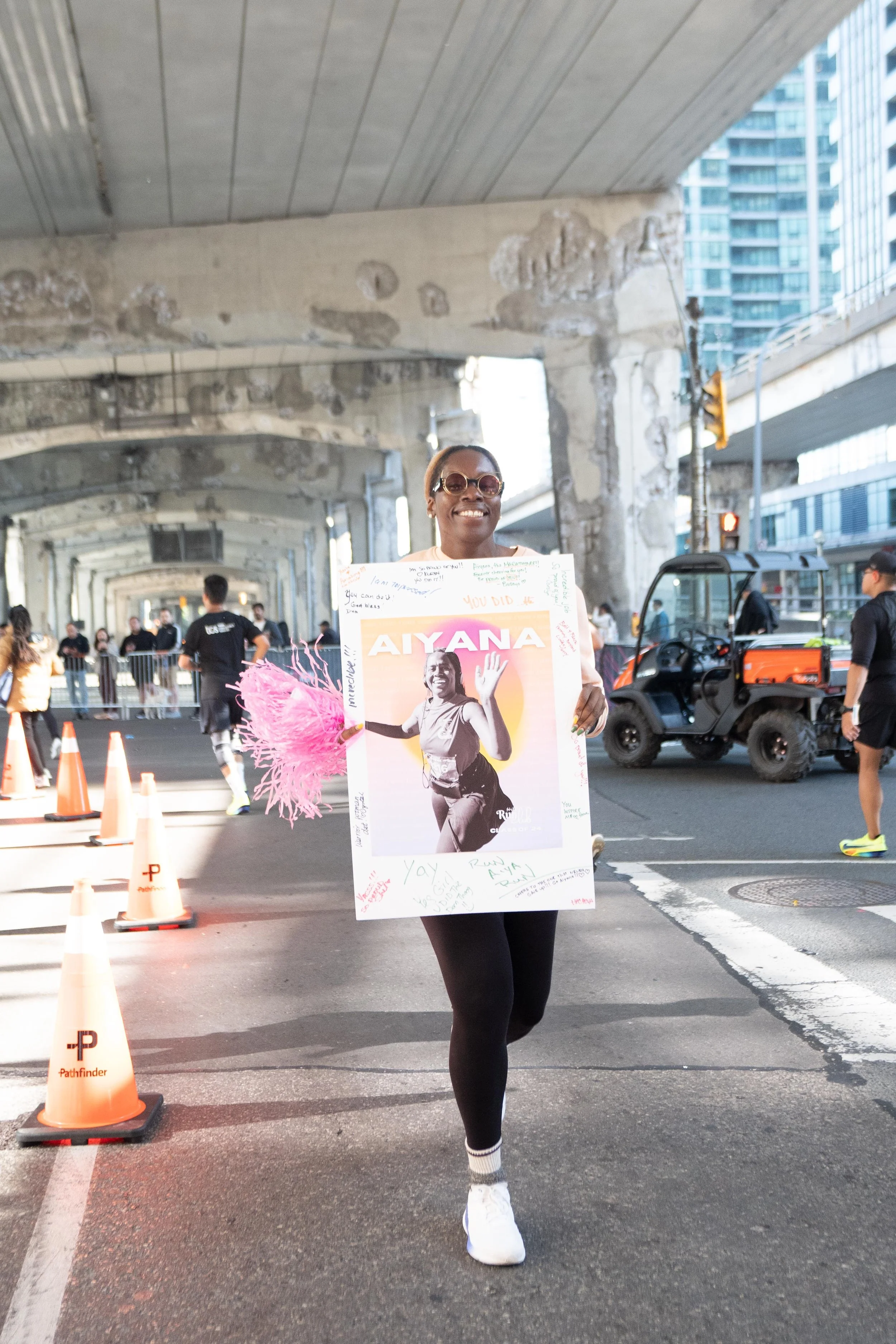 A woman smiling and holding a large poster of herself at a running event under an overpass in an urban setting.