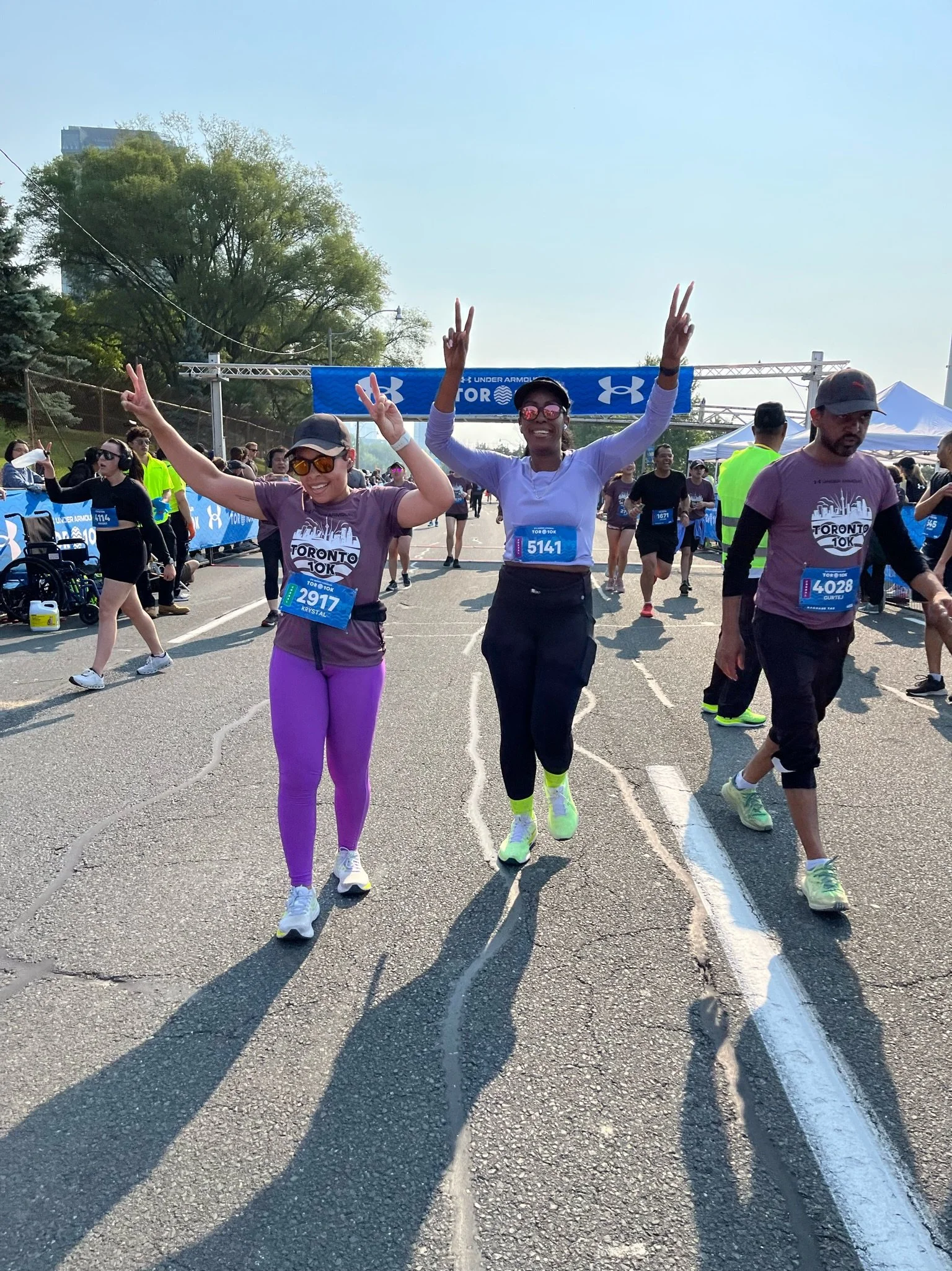Three people celebrating after finishing a race at the Toronto 10K event, standing on the road with race bibs, making peace signs with their hands, surrounded by other runners and event tents.