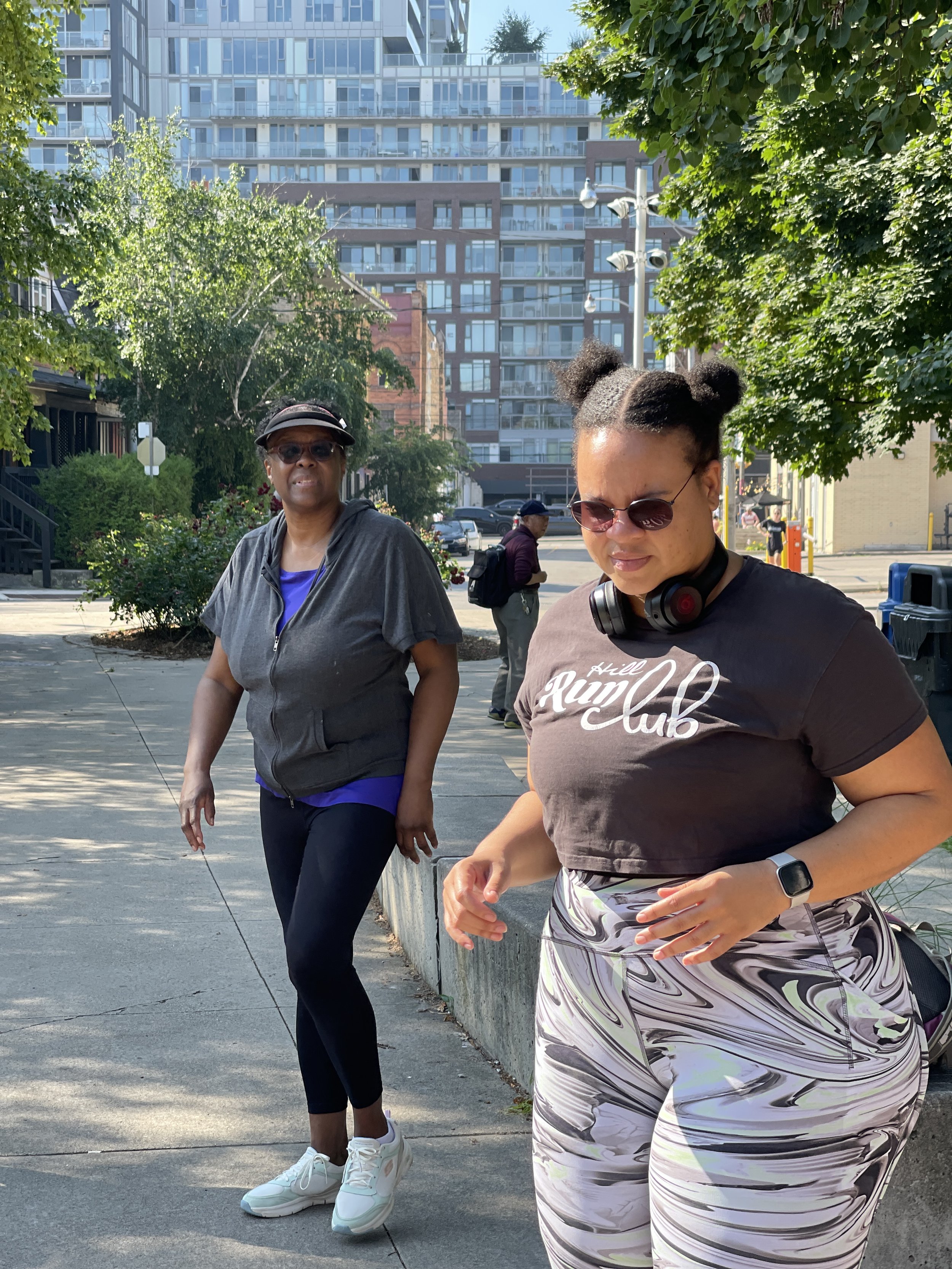 Two women, one older and one younger, are outdoors on a city sidewalk during daytime. The older woman wears sunglasses, a gray zip-up hoodie, black leggings, white sneakers, and a black cap. The younger woman has sunglasses, headphones around her nec