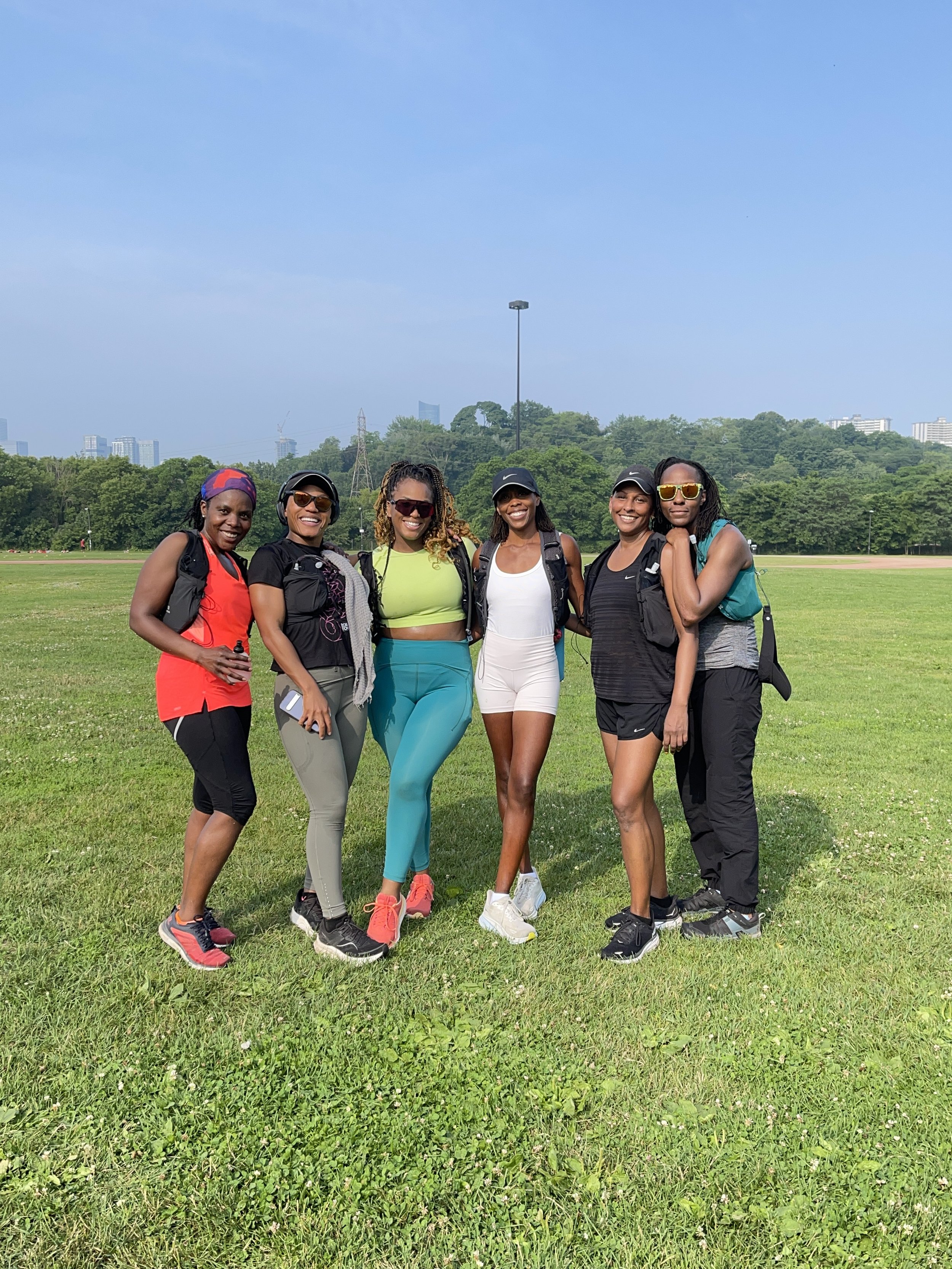 Group of six women in athletic wear standing on grass in park, smiling, with trees and cityscape in background.